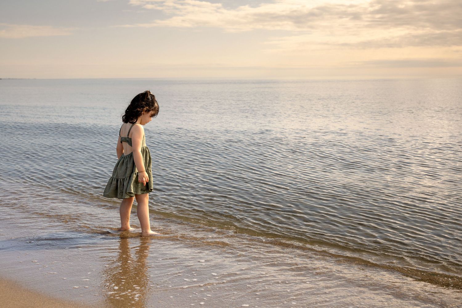 Photographie d'une petite fille en robe à fleurs se tenant pieds nus en bord de mer à Valras plage lors d'une séance photo lifestyle.