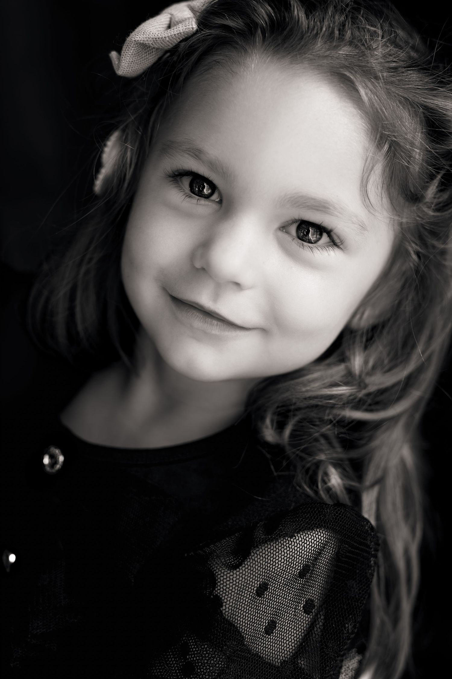 Black and white portrait of a smiling young girl with curly hair and a bow, wearing a dotted dress.