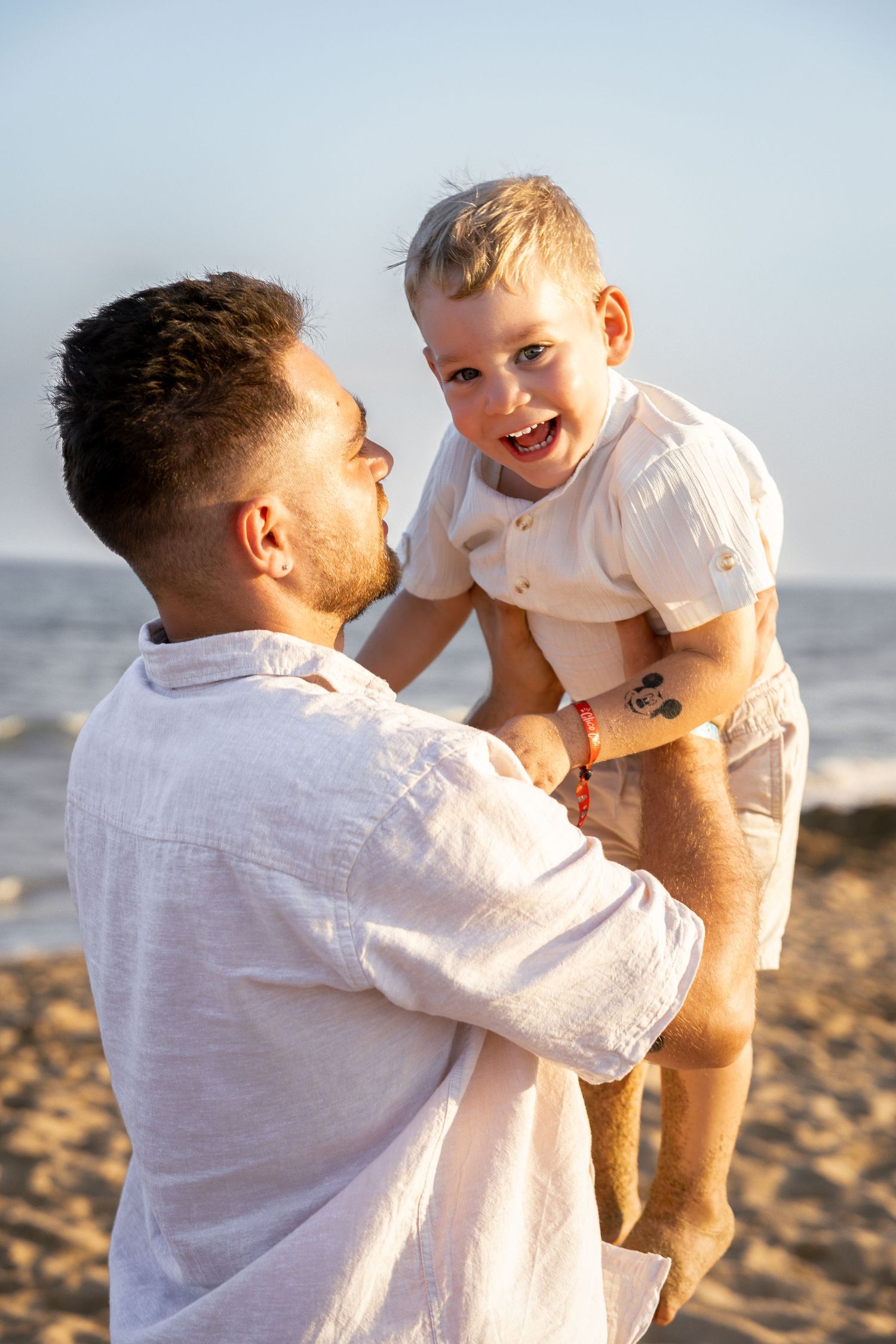 Photo d'un père soulevant un enfant heureux sur une plage ensoleillée, tous deux vêtus de blanc, partageant un moment joyeux dans la mer.