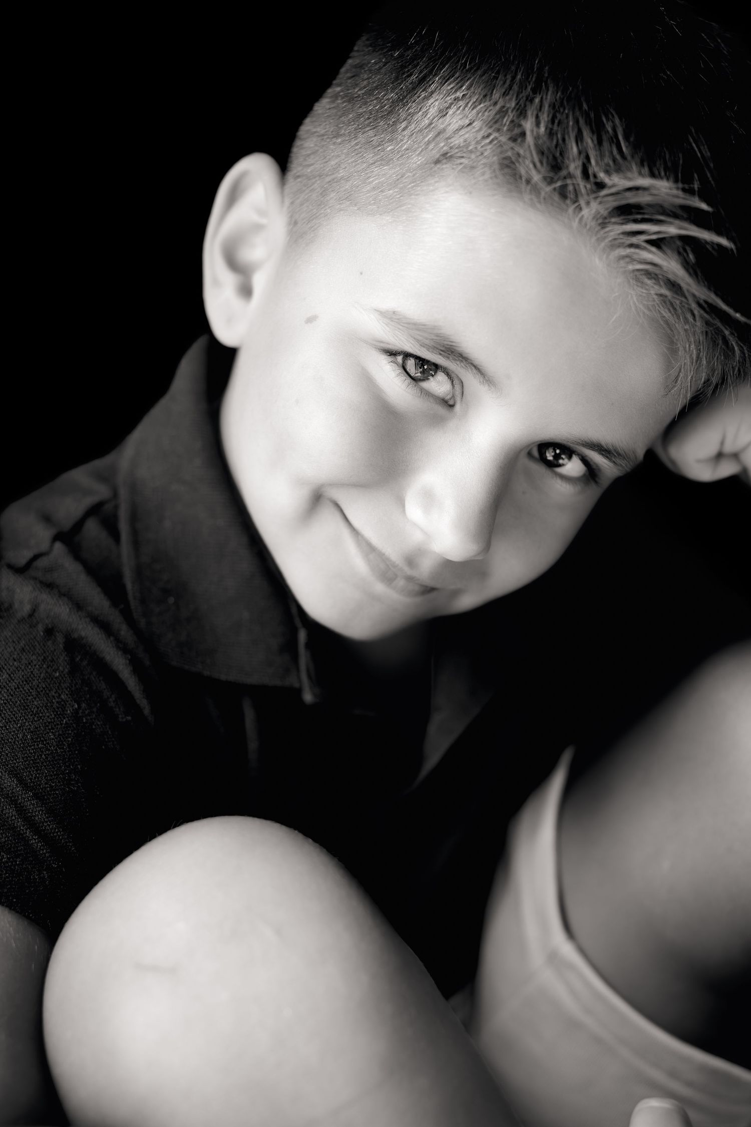 Black and white portrait of a smiling young boy with short hair, wearing a dark polo shirt against a black background.