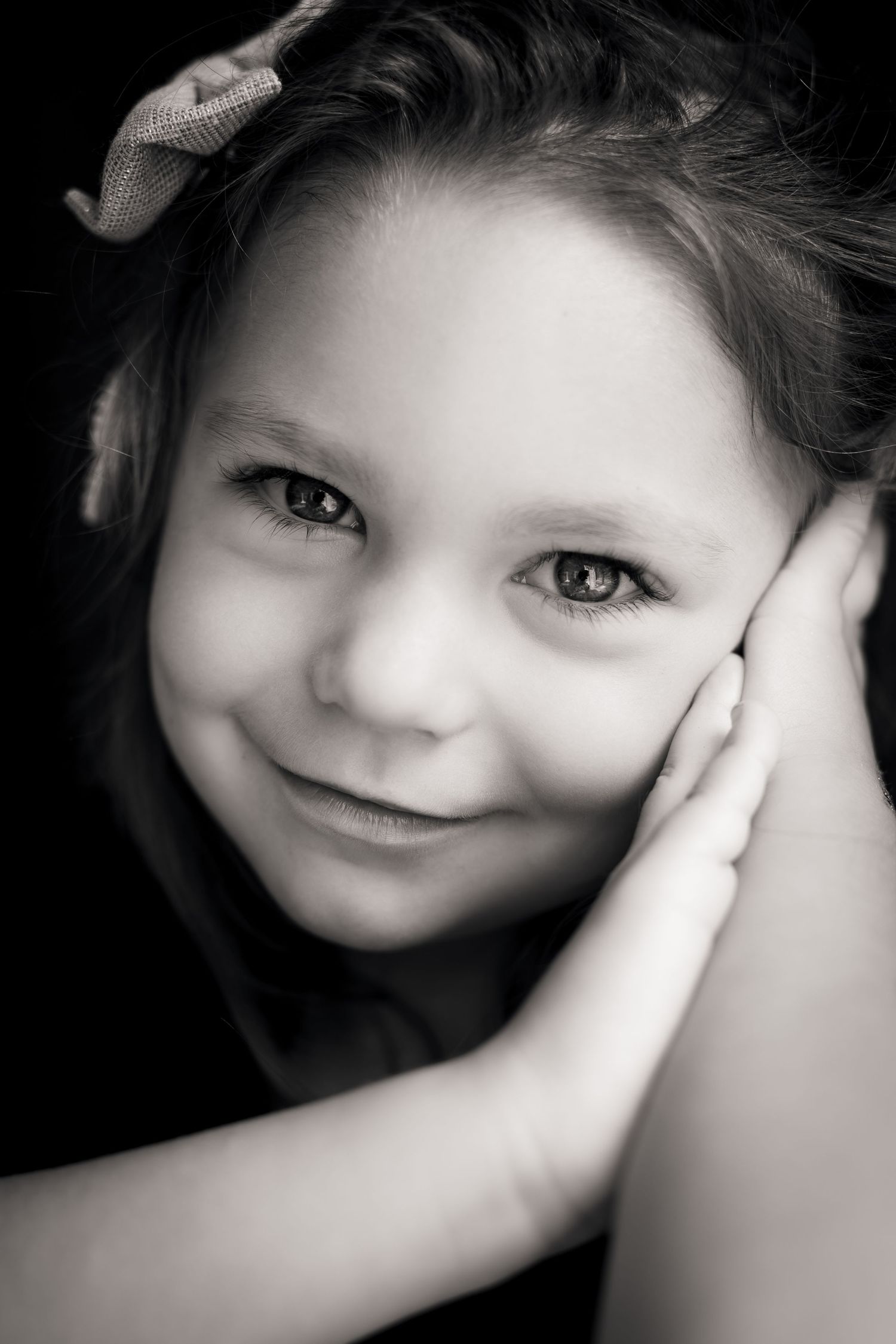 Black and white portrait of a smiling young girl with a bow in her hair, resting her cheek on her hand.