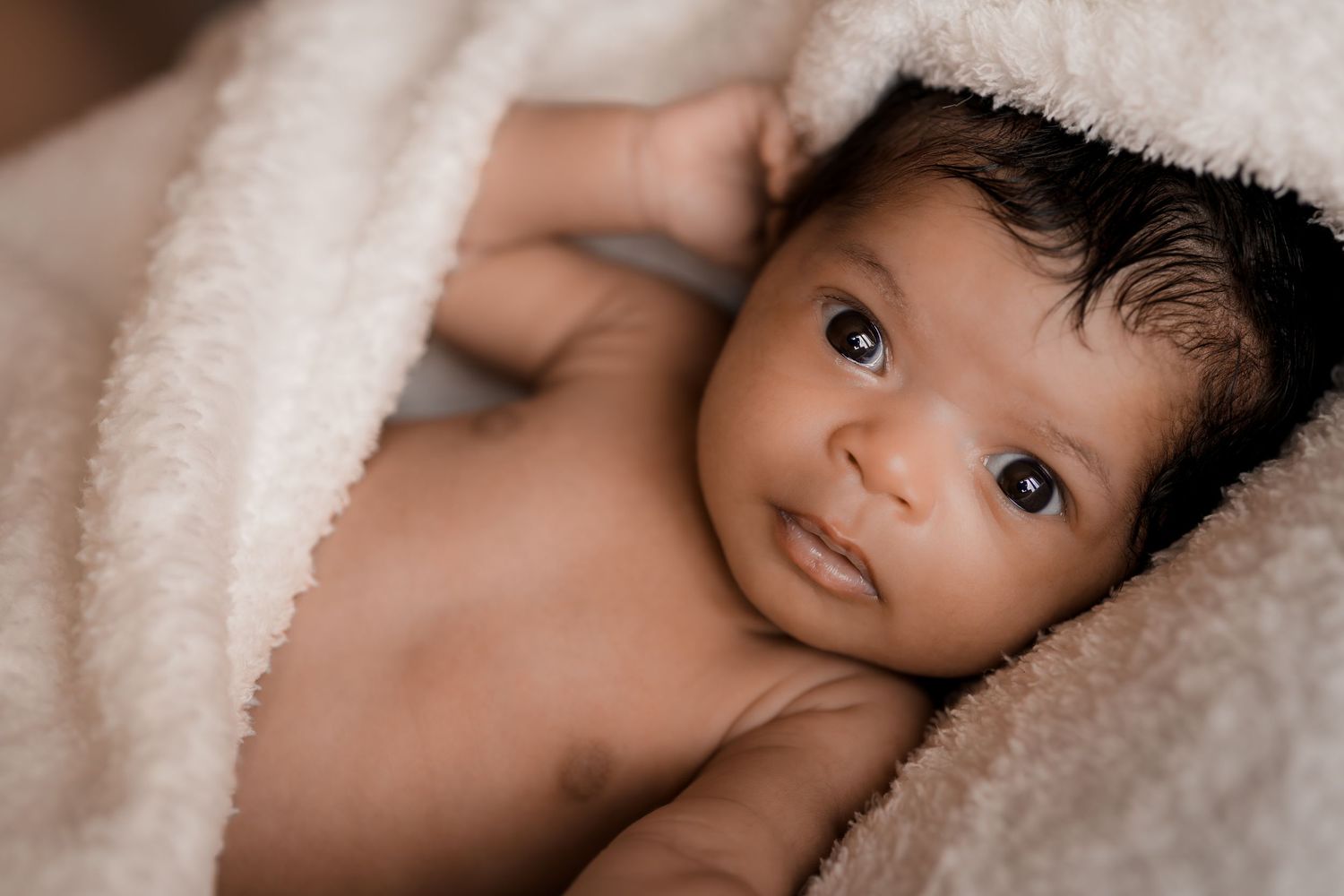 Nouveau-né aux cheveux noirs sortant d'une douce couverture blanche et moelleuse avec des yeux curieux. Photographie naissance Béziers.