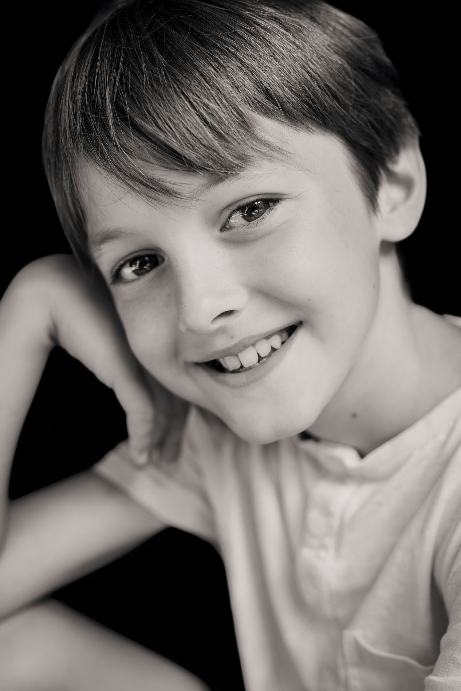 Black and white portrait of a smiling young boy resting his chin on his hand against a dark background.