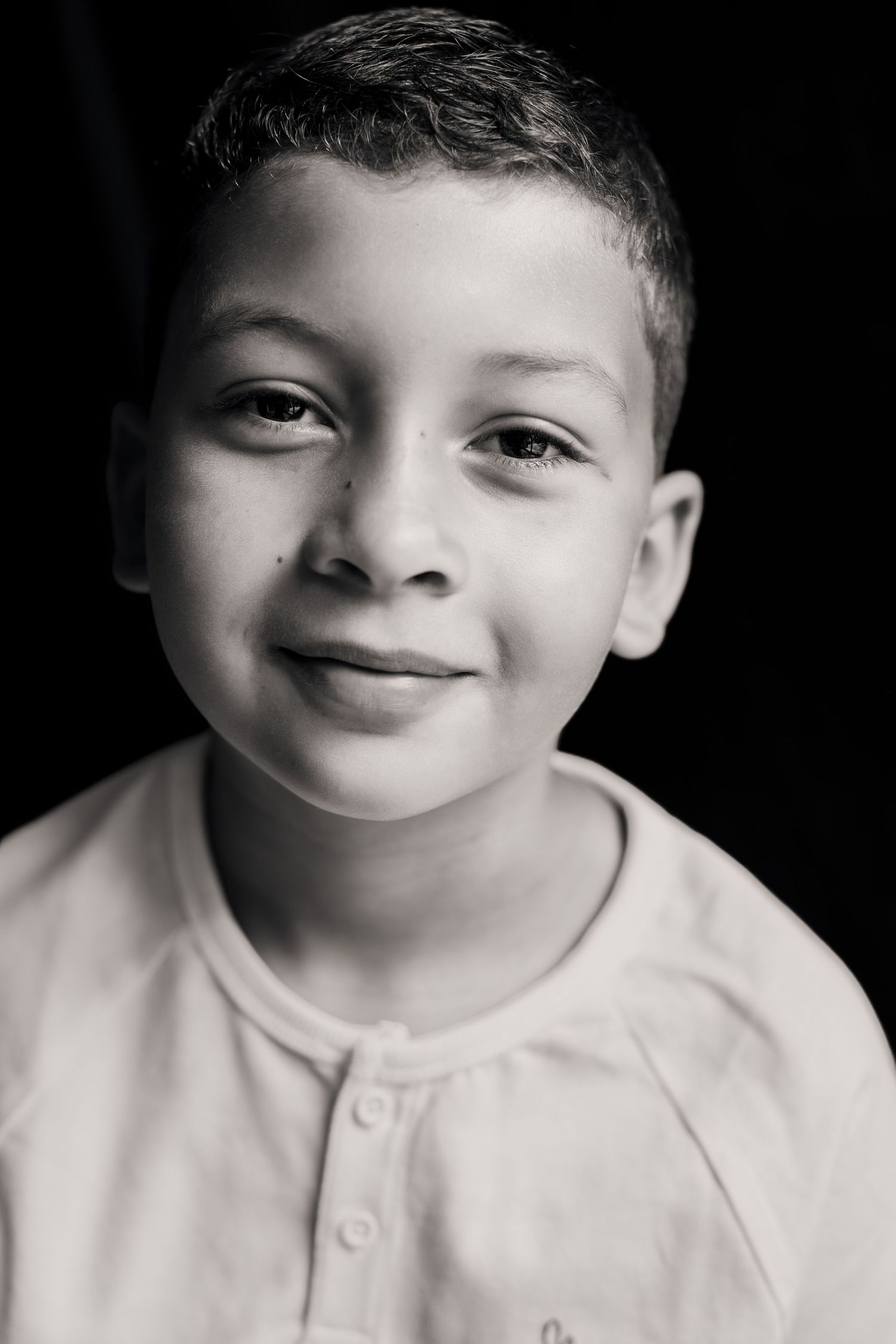 Black and white portrait of a young smiling boy wearing a light-colored shirt against a dark background.