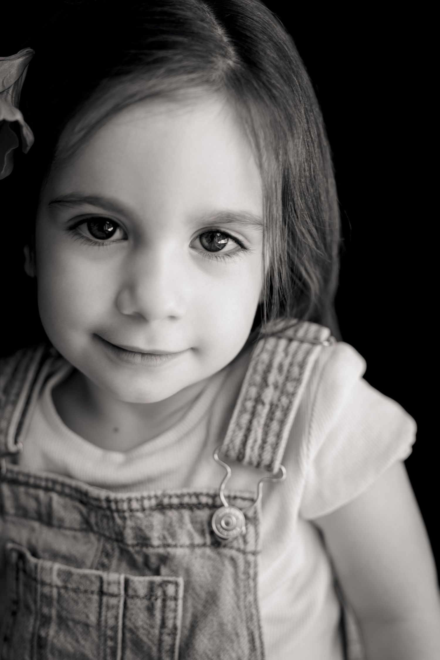 Black and white portrait of a young girl in denim overalls smiling softly against a dark background.