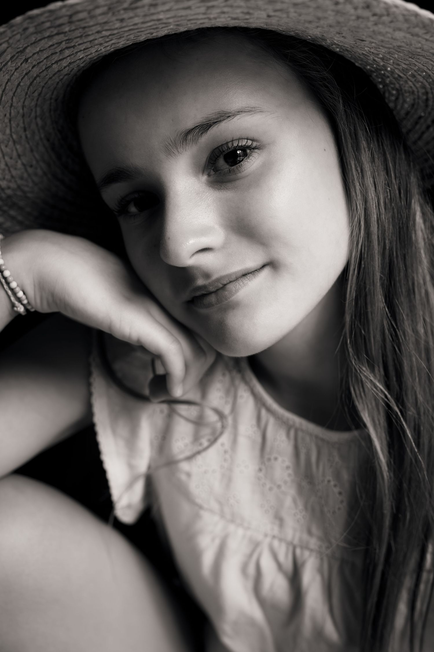 Black and white portrait of a young girl wearing a straw hat, resting her chin on her hand, smiling softly.