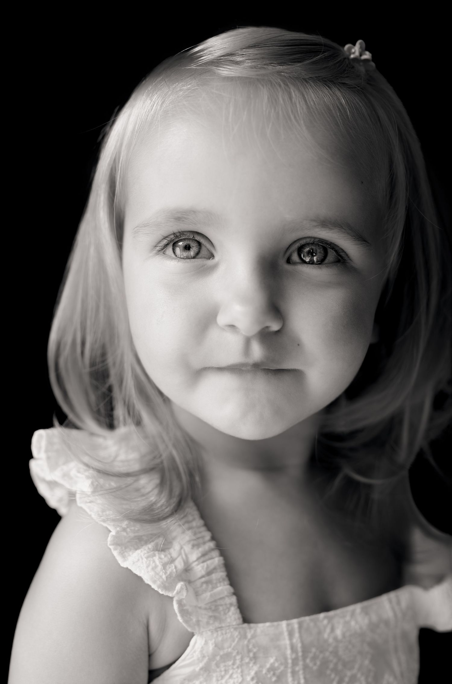 Black and white portrait of a young girl with big eyes and long hair wearing a white ruffled dress.
