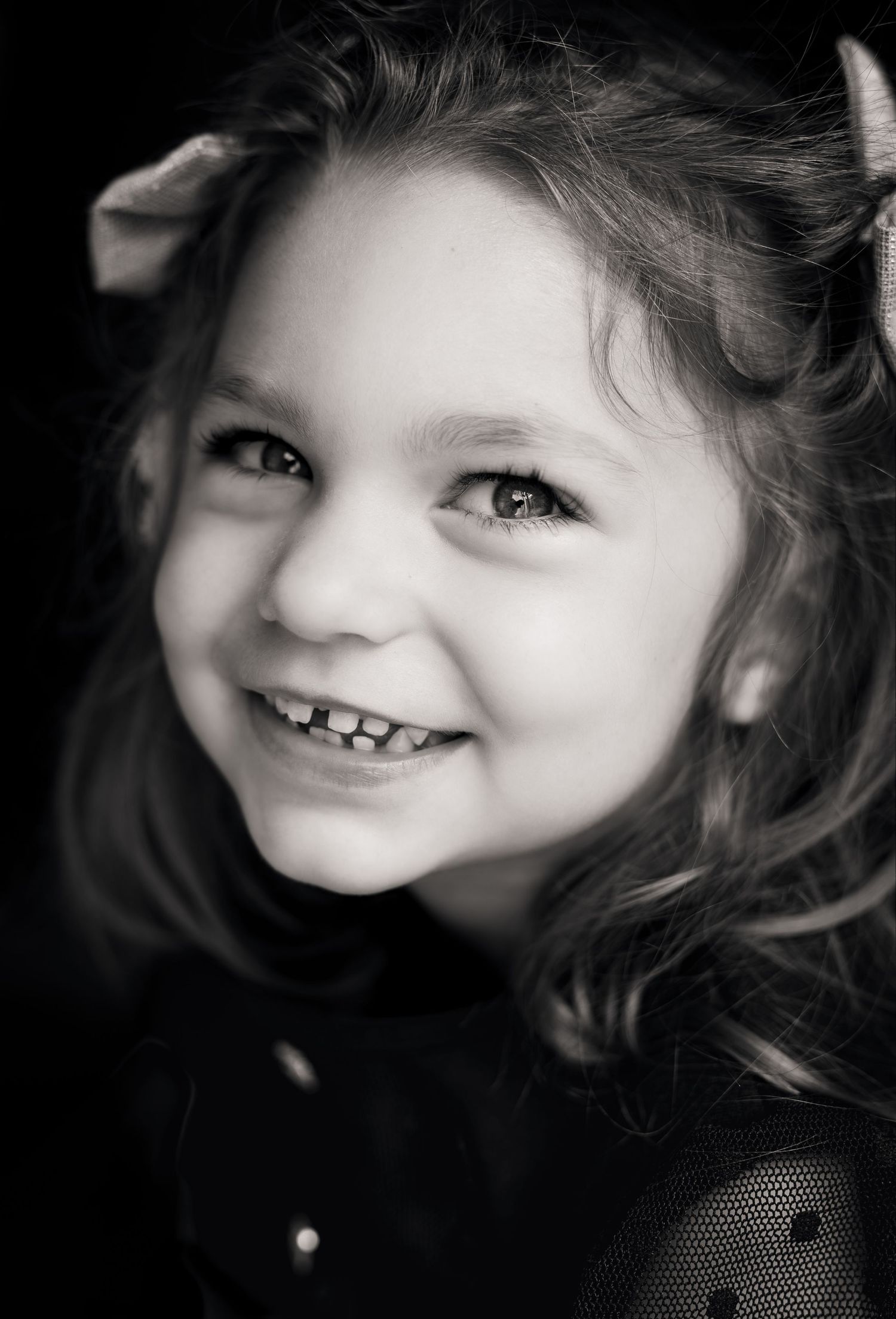 Black and white portrait of a smiling young girl with curly hair and bows, showcasing joy and innocence.