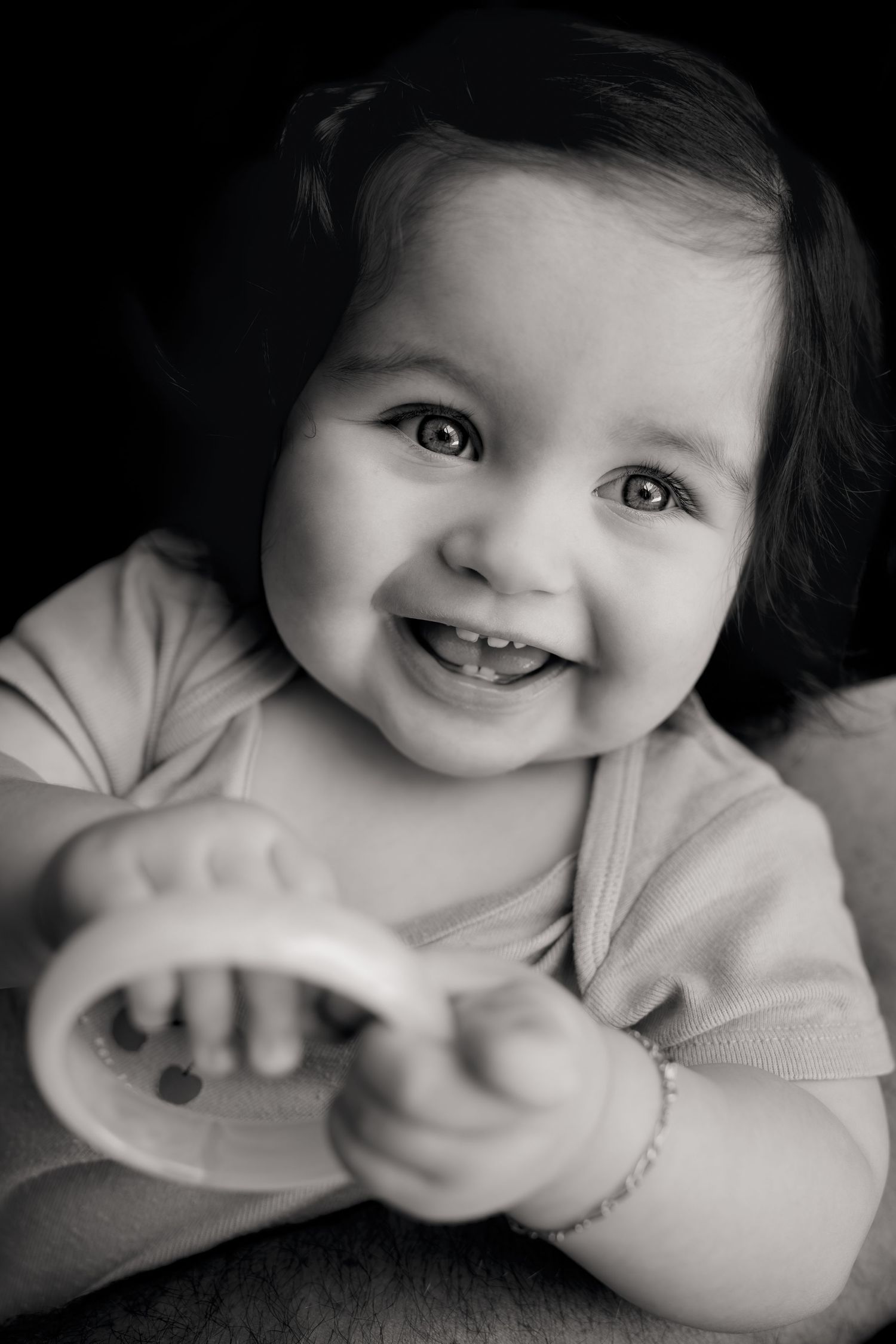 Black and white portrait of a happy smiling baby holding a toy rattle, showing tiny teeth.