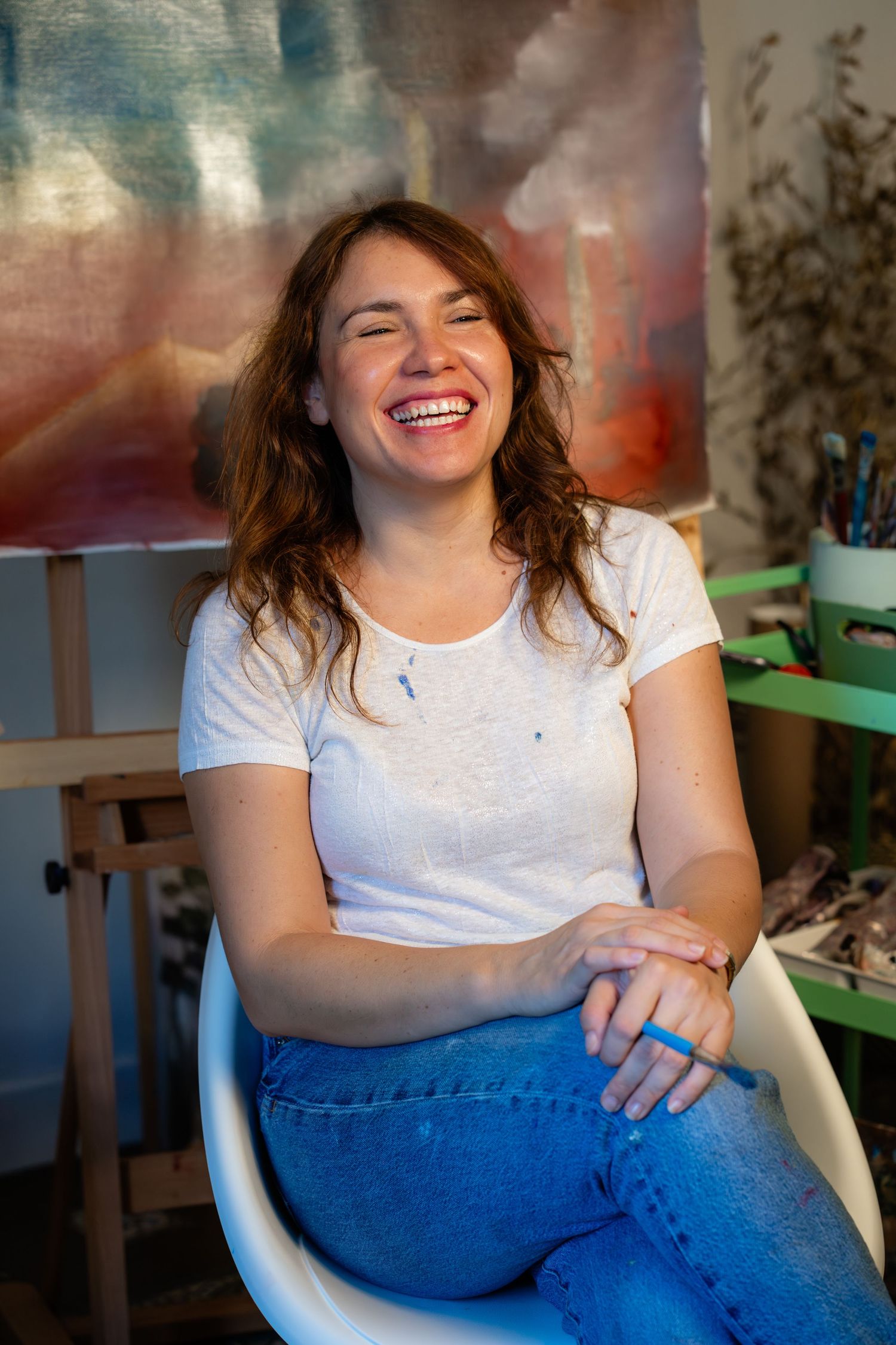 A person in a white t-shirt and blue jeans sits casually in a chair, showing a relaxed and cheerful expression.