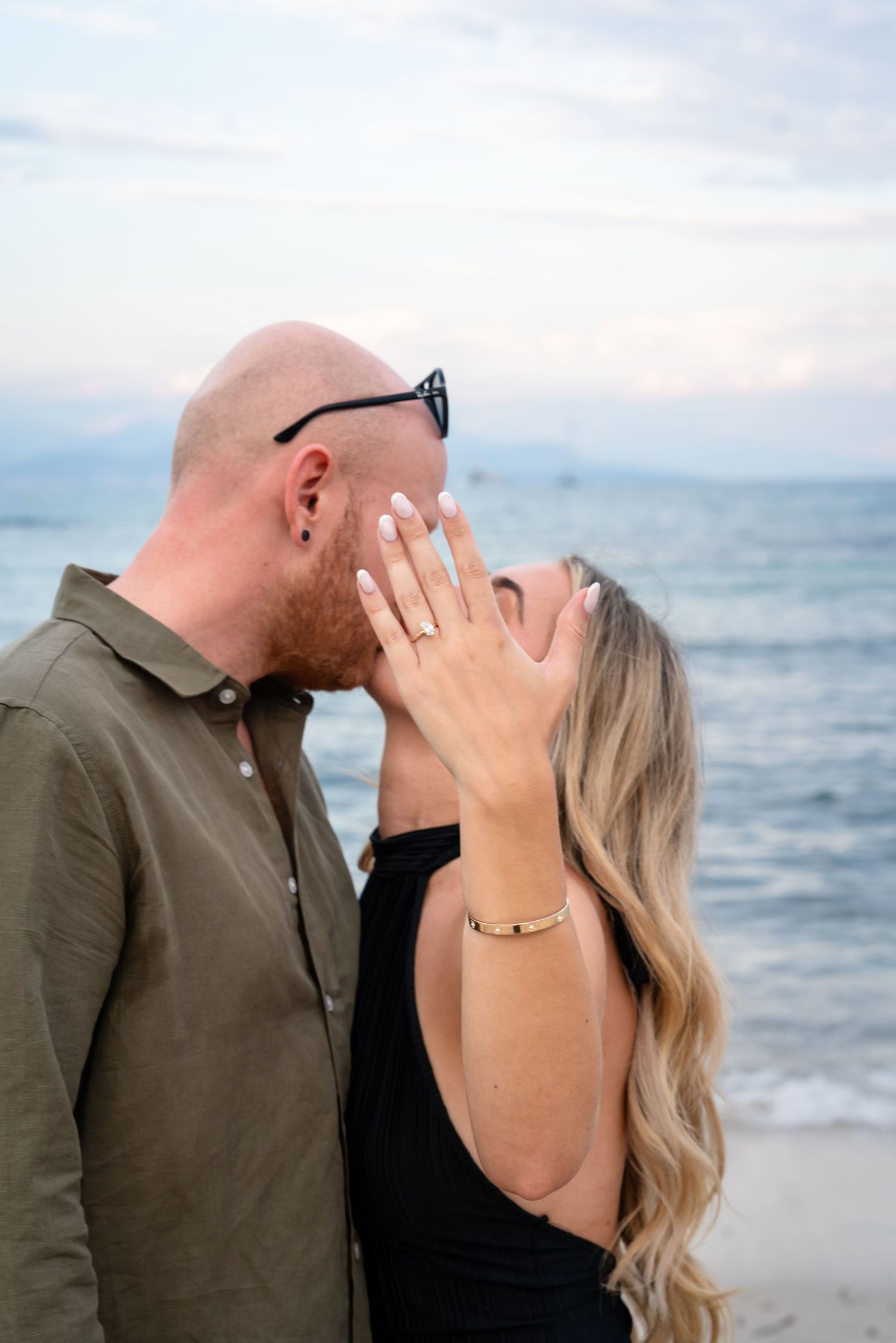 Couple kissing on beach at sunset, woman showing engagement ring with ocean backdrop.