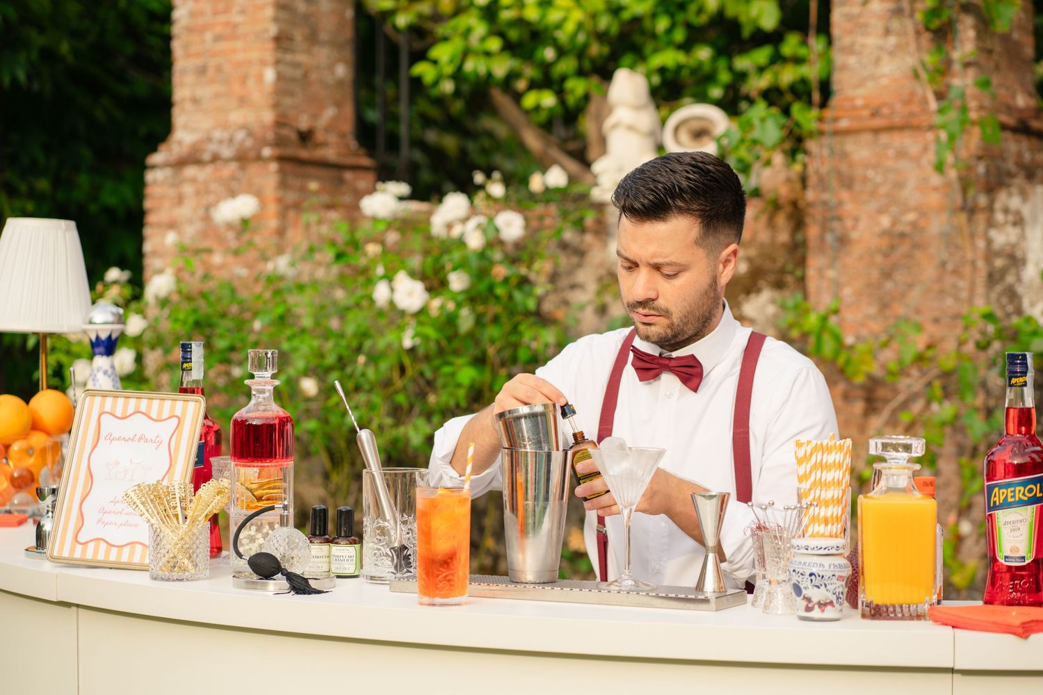 Bartender in suspenders and bow tie mixing cocktails at an outdoor bar.