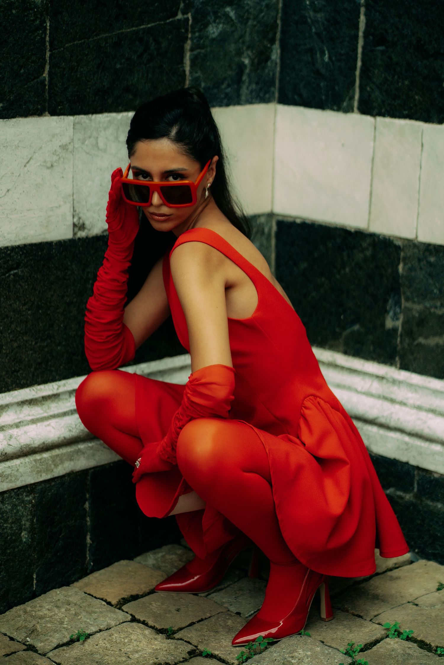 Dramatic fashion shoot with model in red dress and gloves crouching against dark tiles.