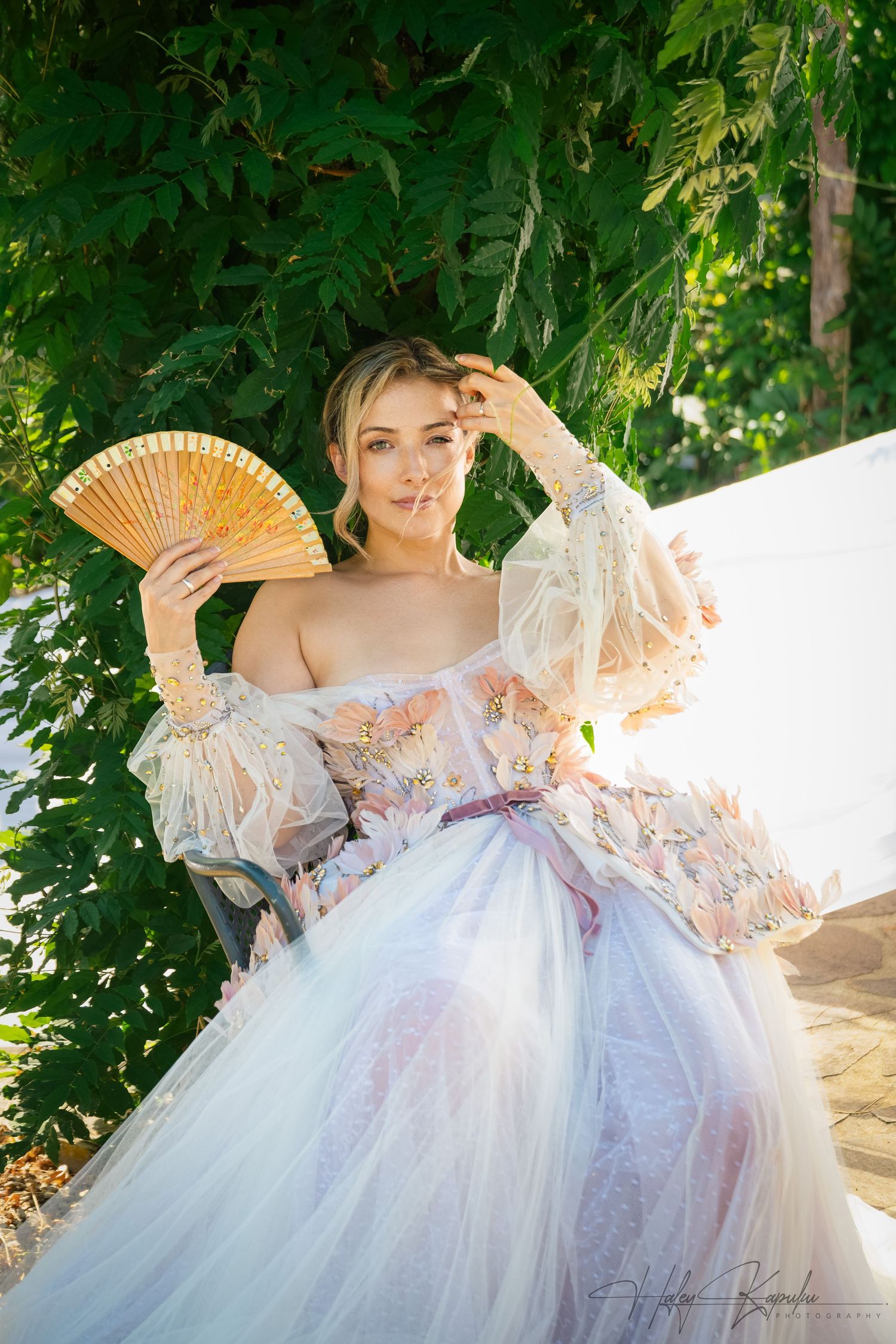 Ethereal white lace dress with floral details photographed in a garden setting with a decorative fan.
