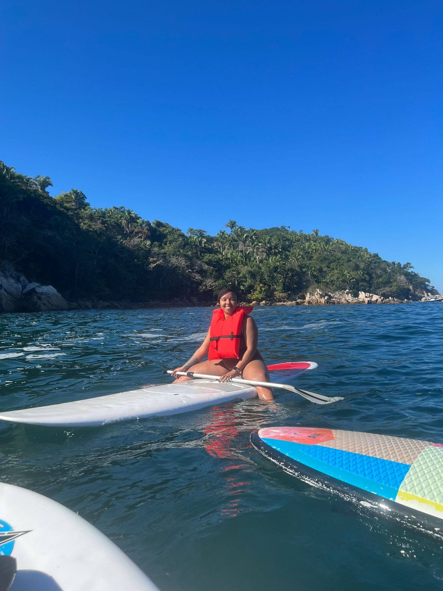 Person sitting on paddleboard in blue waters near rocky coastline.