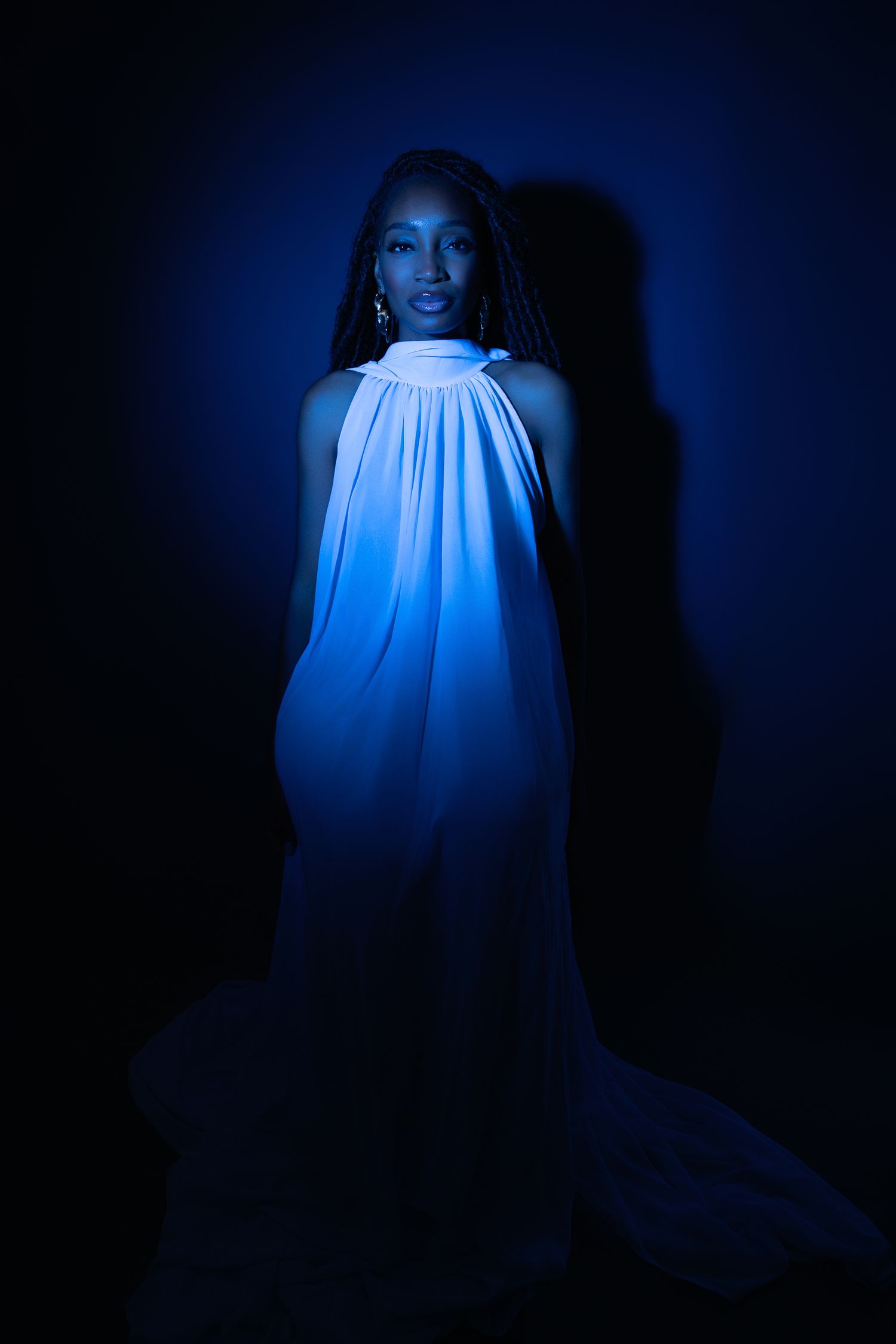 A woman in a flowing white halter dress illuminated by dramatic blue lighting against a dark background.