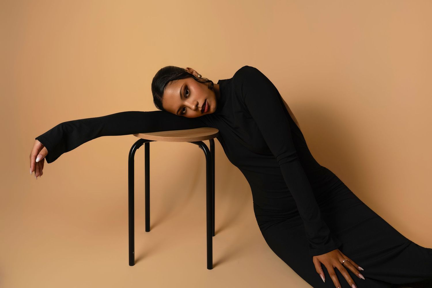A woman in a black outfit drapes elegantly over a wooden stool against a beige background.