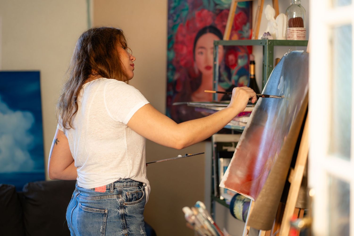 Artist in white t-shirt and jeans works on a colorful painting at an easel in their studio space.