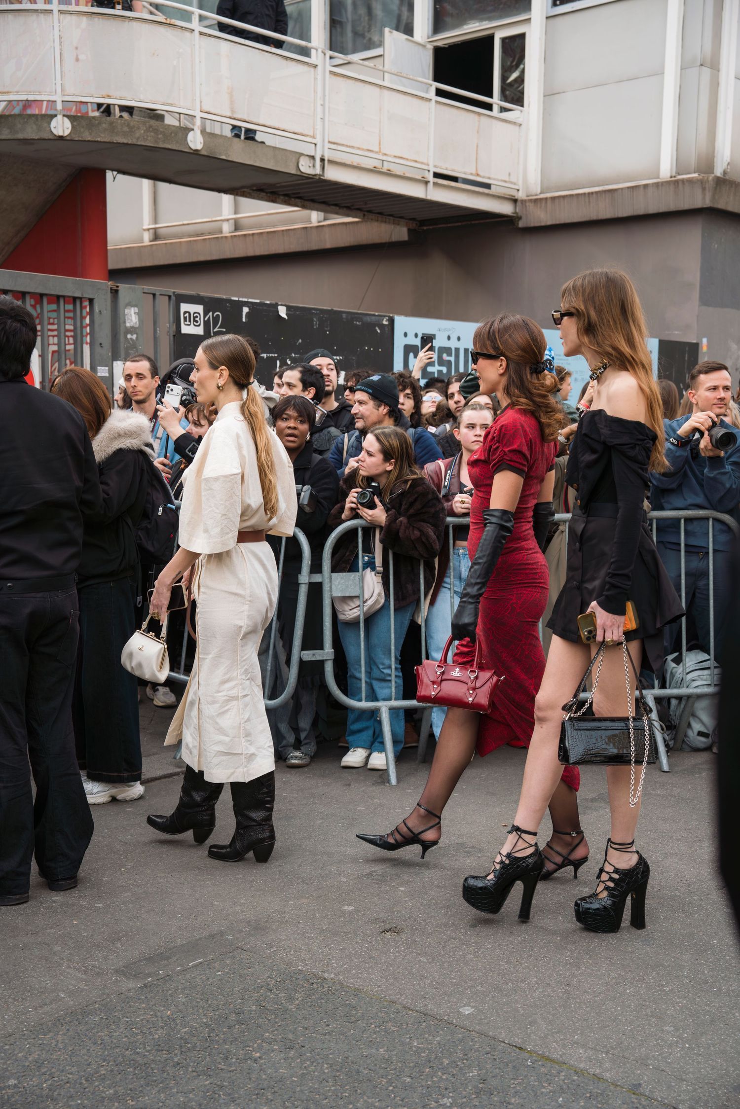 Fashion week attendees in stylish outfits wait behind barriers at an outdoor event venue.