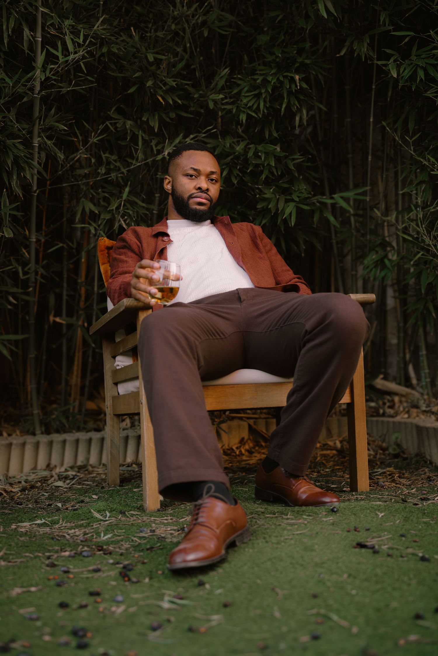 Person lounging in wooden chair wearing brown jacket and boots against bamboo backdrop.