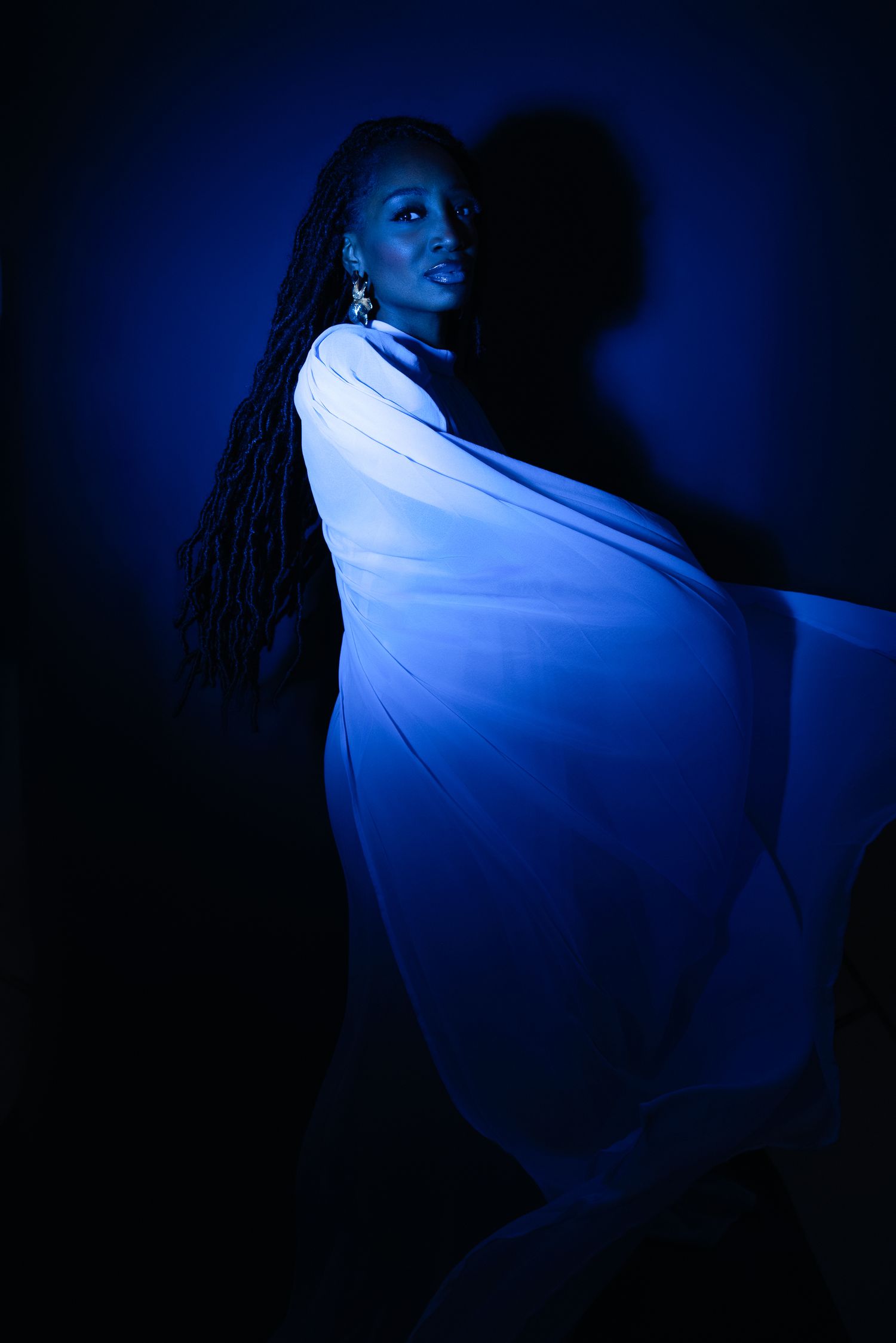 A woman draped in flowing white fabric poses dramatically under deep blue lighting against a dark background.