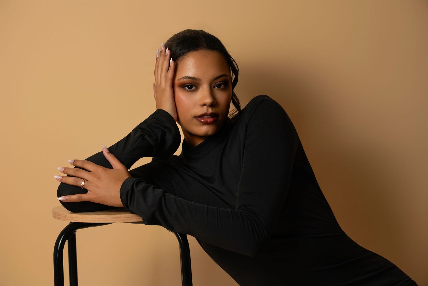 Model wearing a black turtleneck sweater poses elegantly on a chair against a warm beige backdrop.