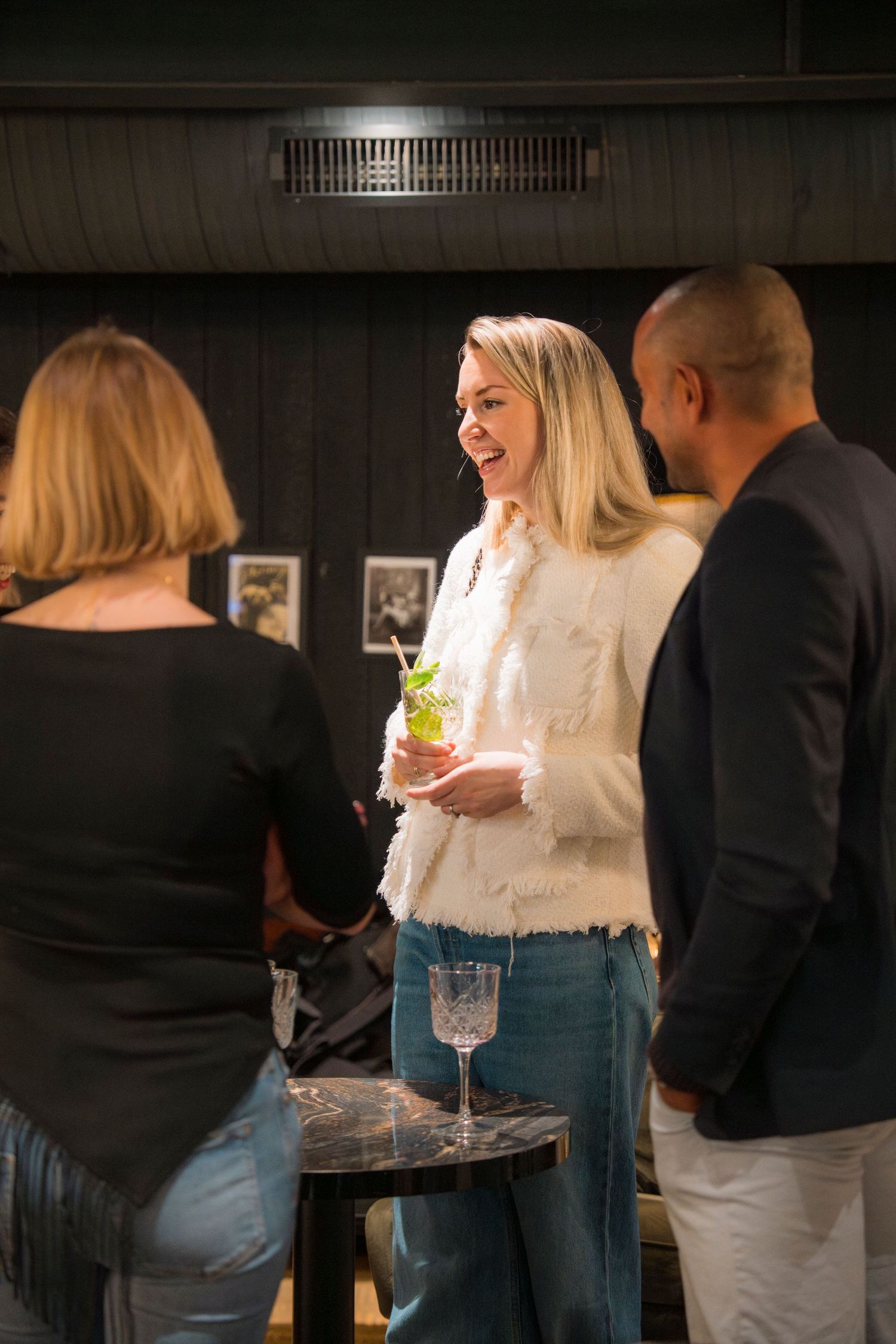People gathered at a dimly lit bar socializing and holding drinks in a casual evening setting.