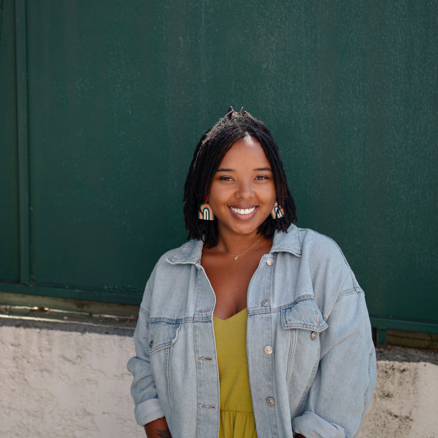 Casual portrait of the owner and photographer in  denim jacket and yellow top against green background.