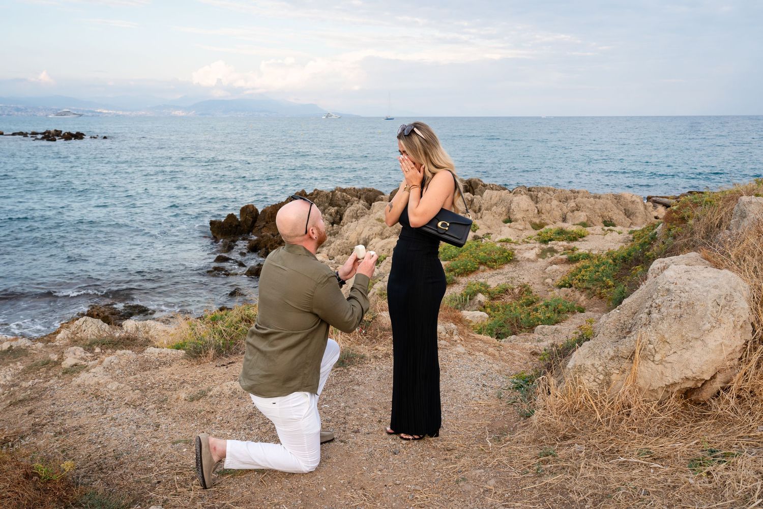 Man proposing to woman in black dress by rocky seaside at sunset, romantic beach marriage proposal scene.