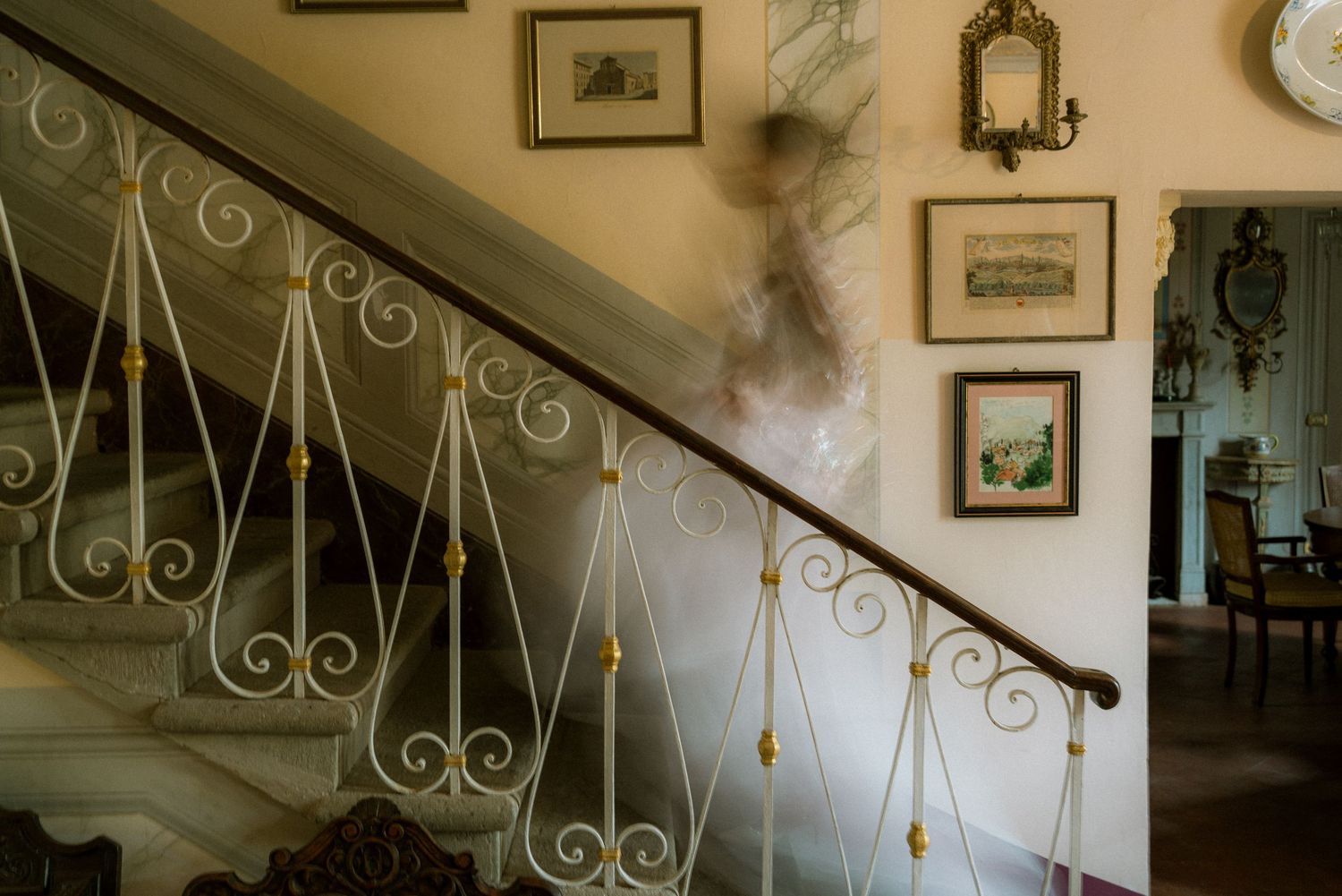 Blurred figure descending an elegant staircase with ornate white iron railings and framed artwork on cream walls.