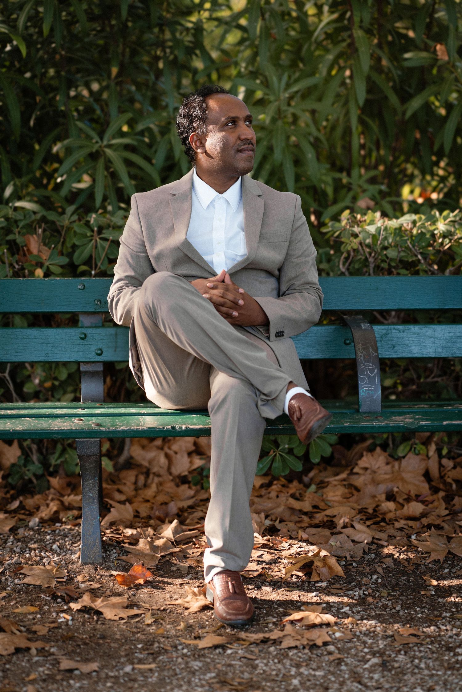 A person in a light grey suit sits thoughtfully on a green park bench surrounded by fallen leaves. Taken at a park in Cannes, France