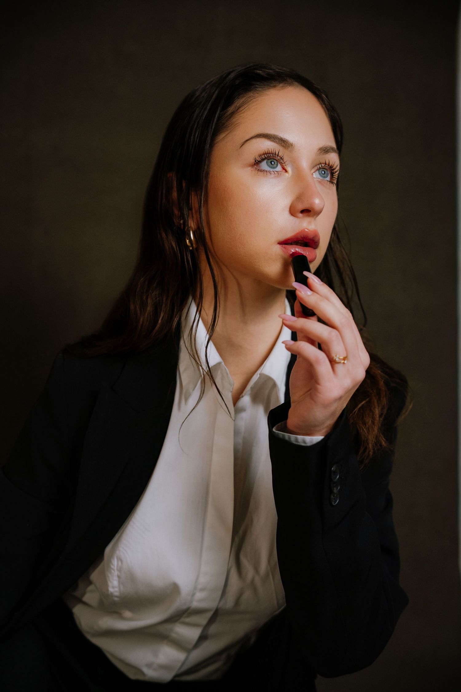 Professional beauty portrait of someone in business attire with dark hair applying lipstick against a moody dark background.