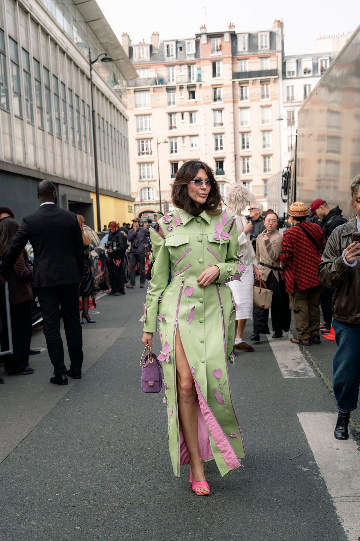 A person in a light green coat walks through a crowded street with historic European buildings in the background.