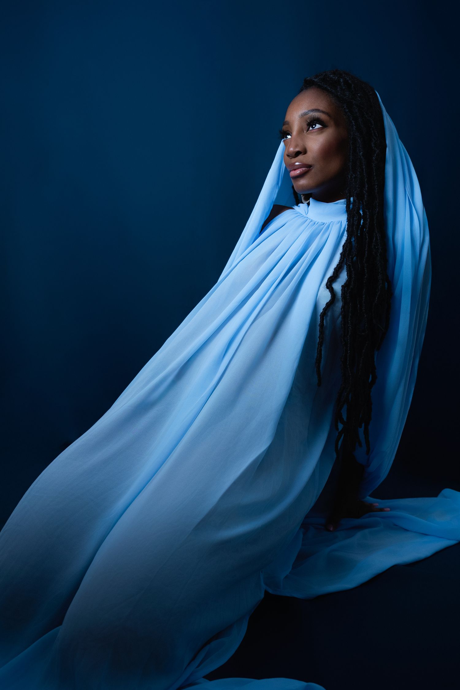 A Black woman with long braids draped in flowing light blue fabric poses against a dark blue background.