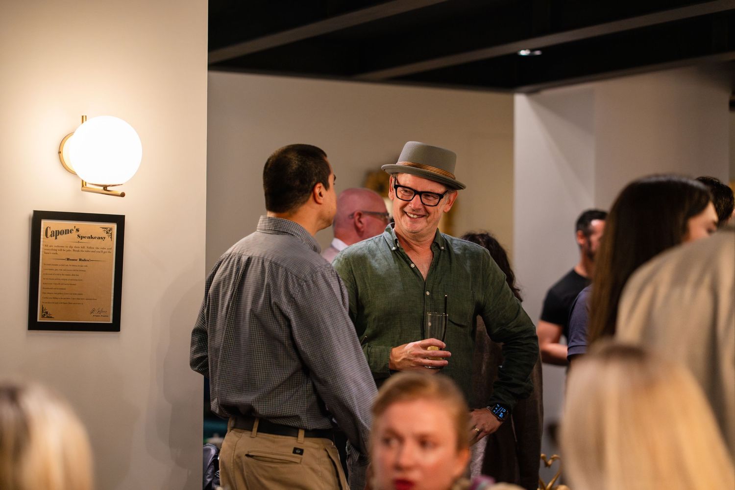 A group of people gather in an art gallery space with framed artwork visible on the walls.