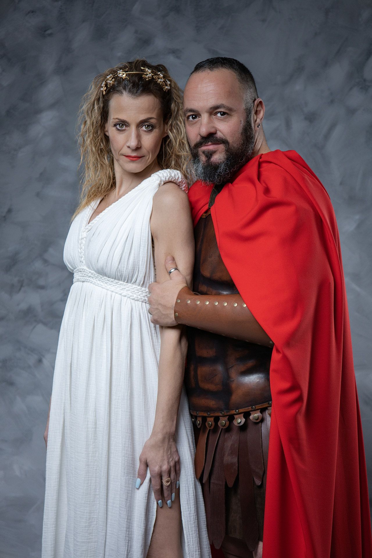A couple in romantic poses wearing contrasting white dress and red cape with leather armor against a gray backdrop.