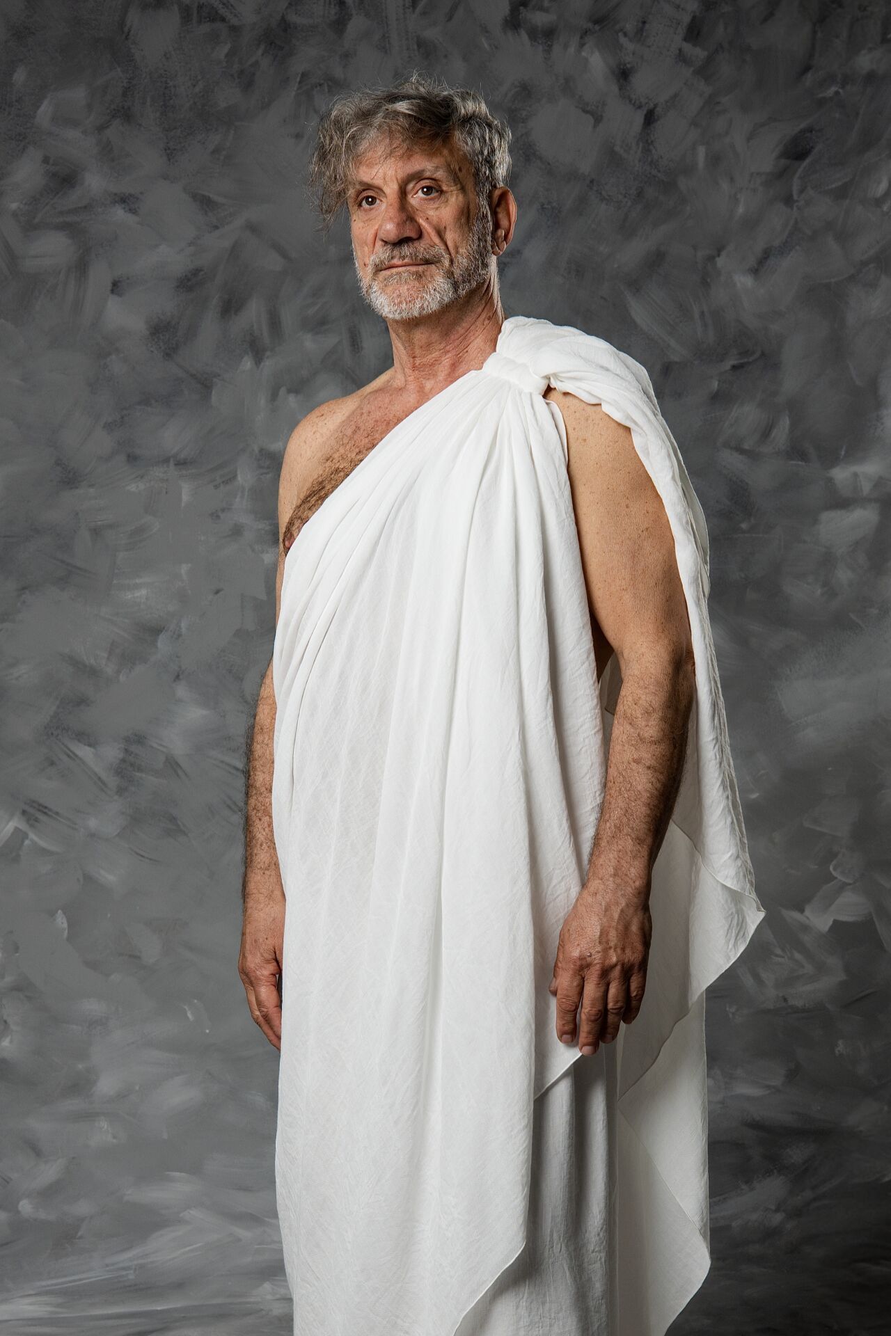 Elderly man in white Roman toga poses against gray textured background in studio portrait.