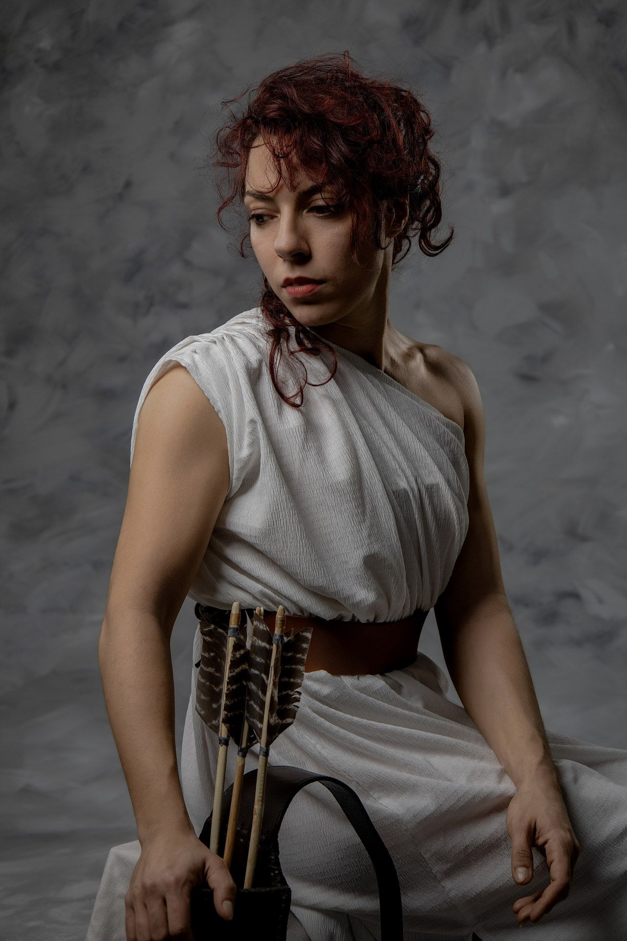 Red-haired woman in white Greek toga dress with leather corset and arrows, posing dramatically against gray background.
