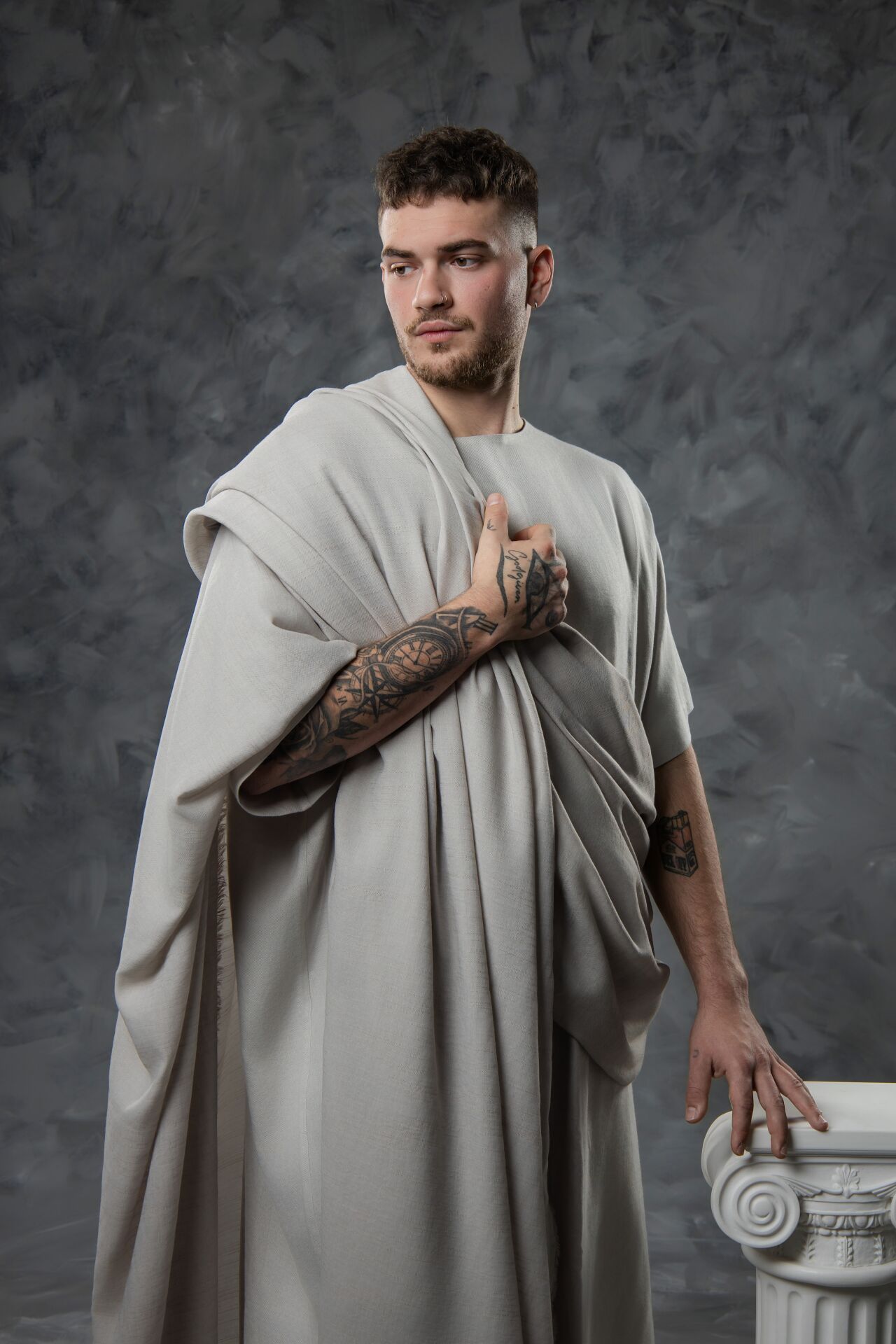 A tattooed man in a Roman toga poses beside a white column against a gray textured background.