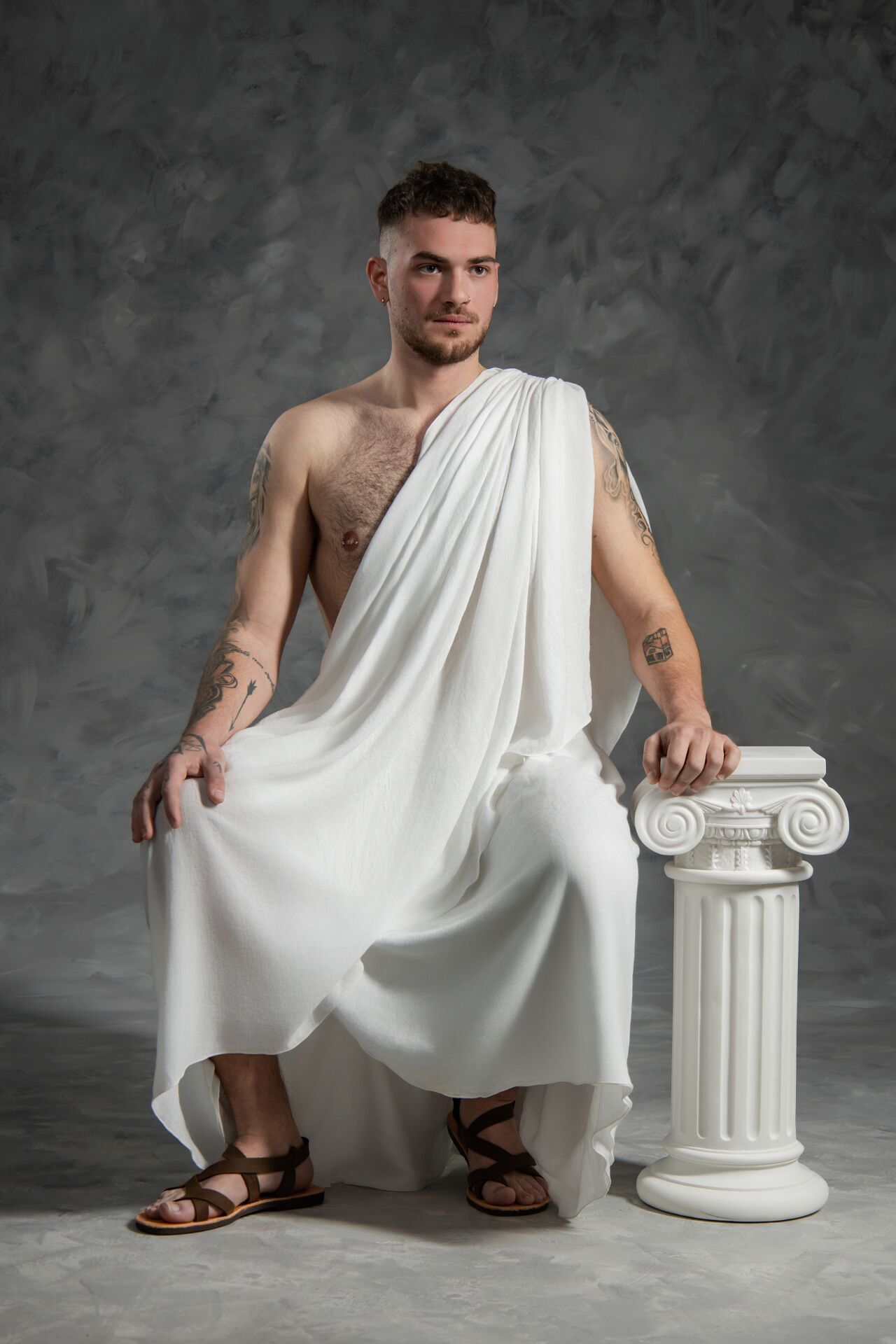 A person in a white toga-style garment poses with a decorative white column in a studio setting against a gray backdrop.