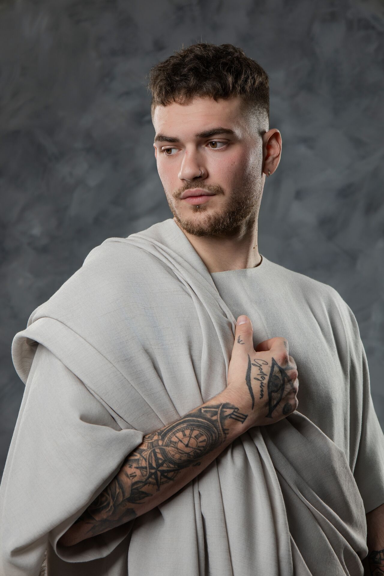 Portrait of someone wearing a light gray draped fabric with visible tattoo sleeve artwork on their arm.