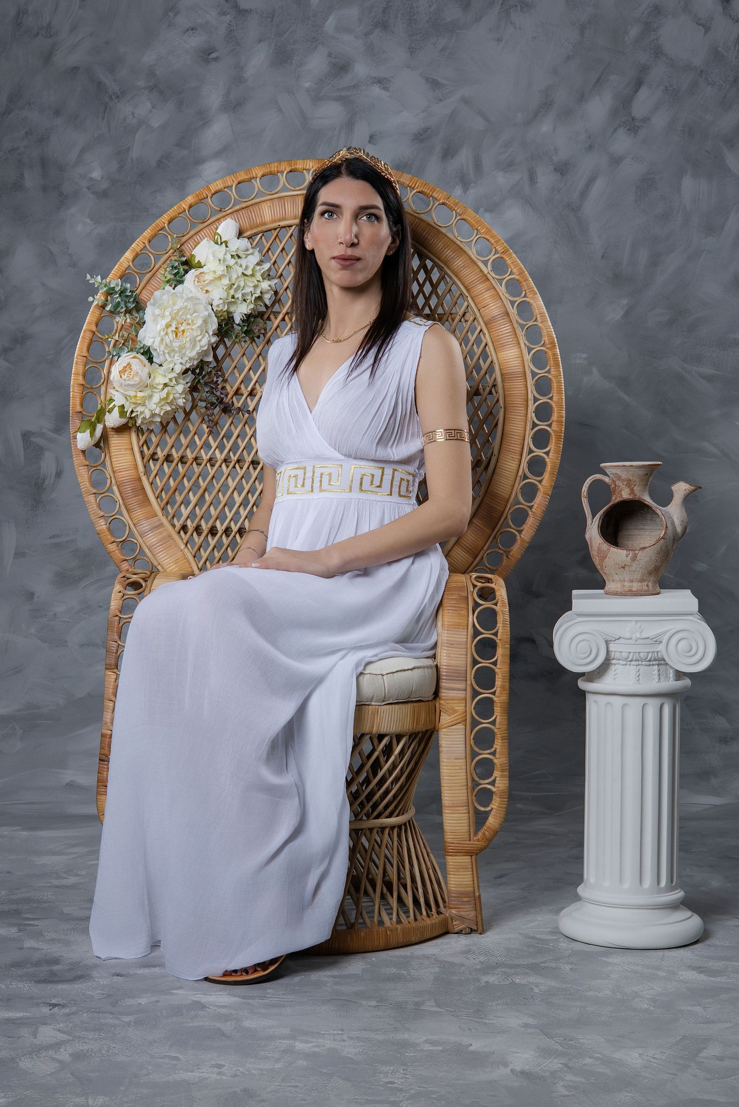 Woman in white Greek goddess dress sits in wicker peacock chair holding flowers, beside a classical column with clay vase.