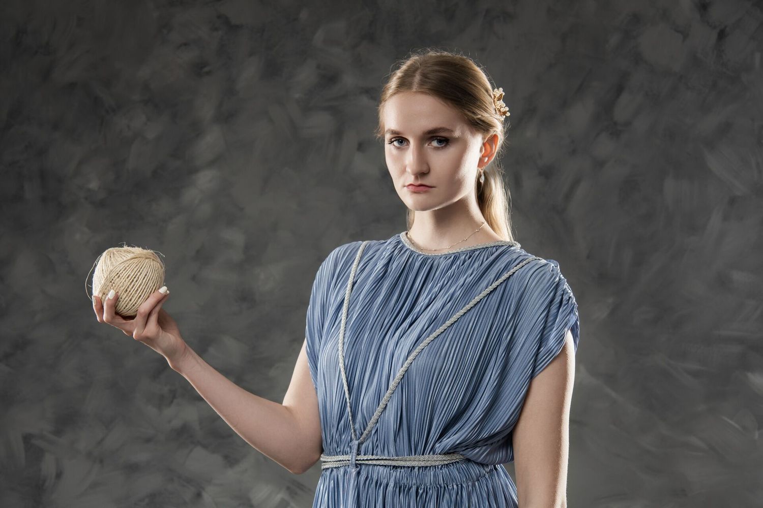 Young woman in blue dress holding a small woven ball, posing against gray studio background.