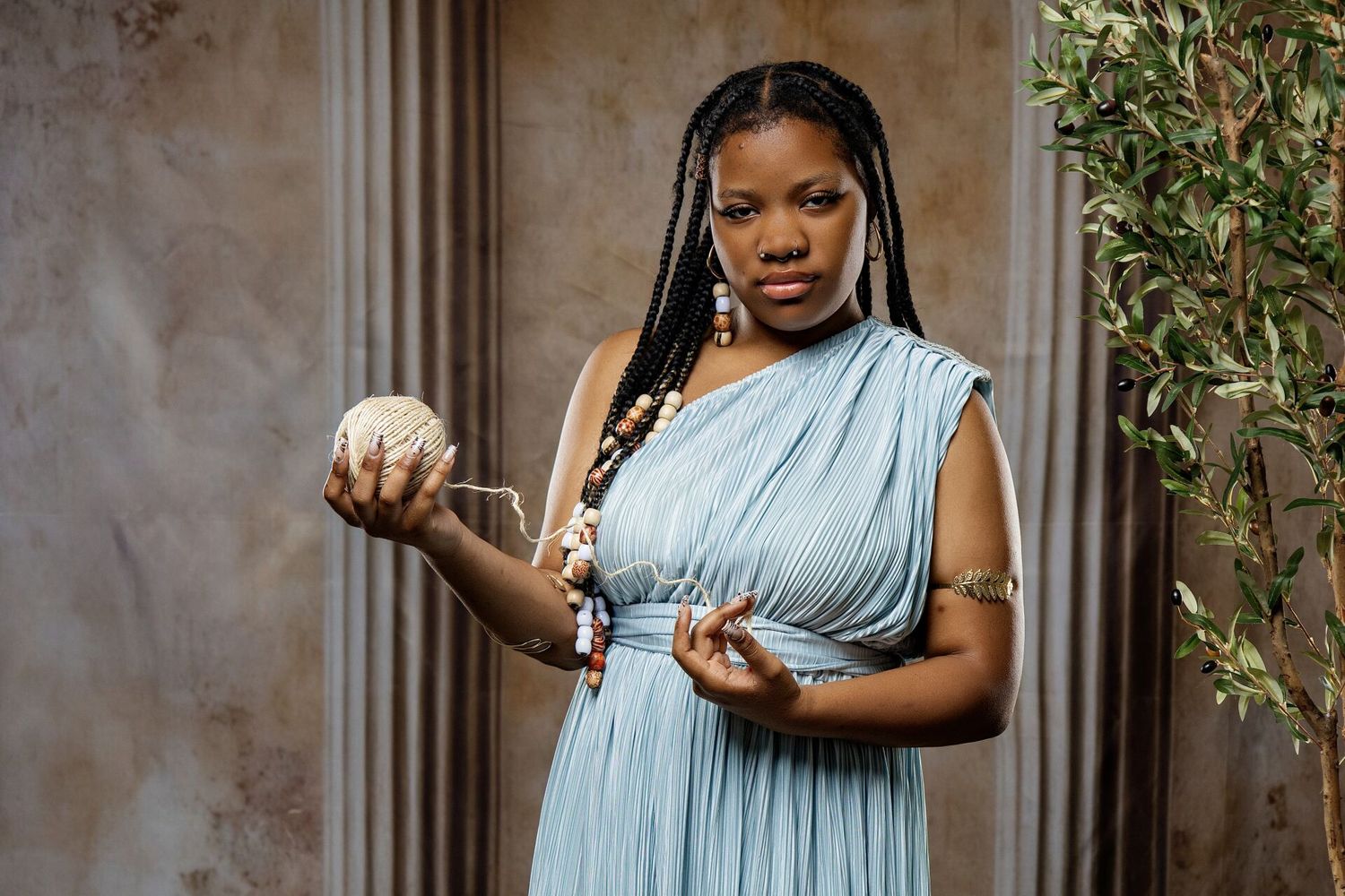 Woman in blue grecian dress holding a decorative object, standing near a column and green plant.