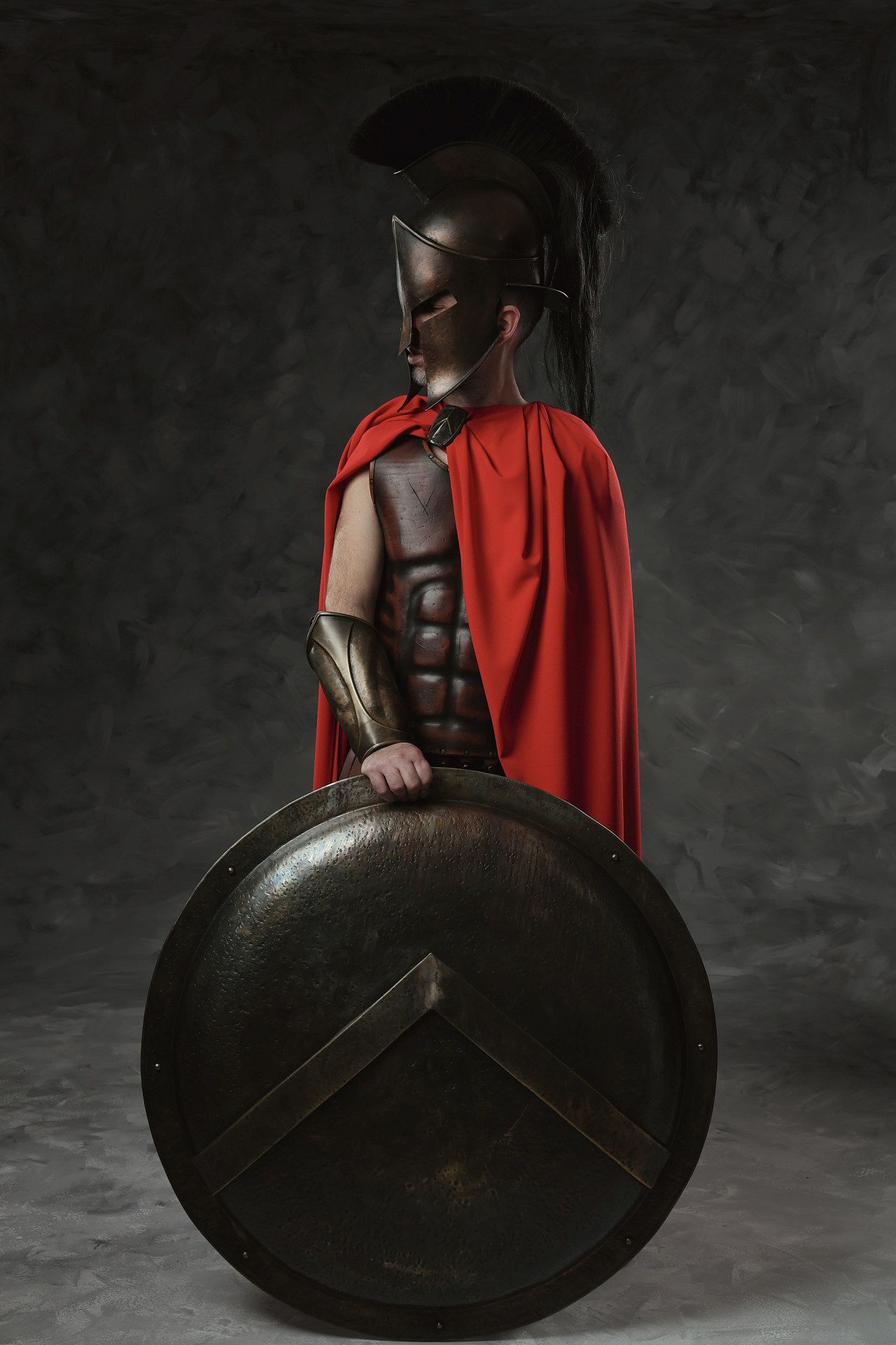 Ancient Greek warrior studio photoshoot of muscular figure in red cape holding large round shield, dramatic portrait by GreekClick, Athens