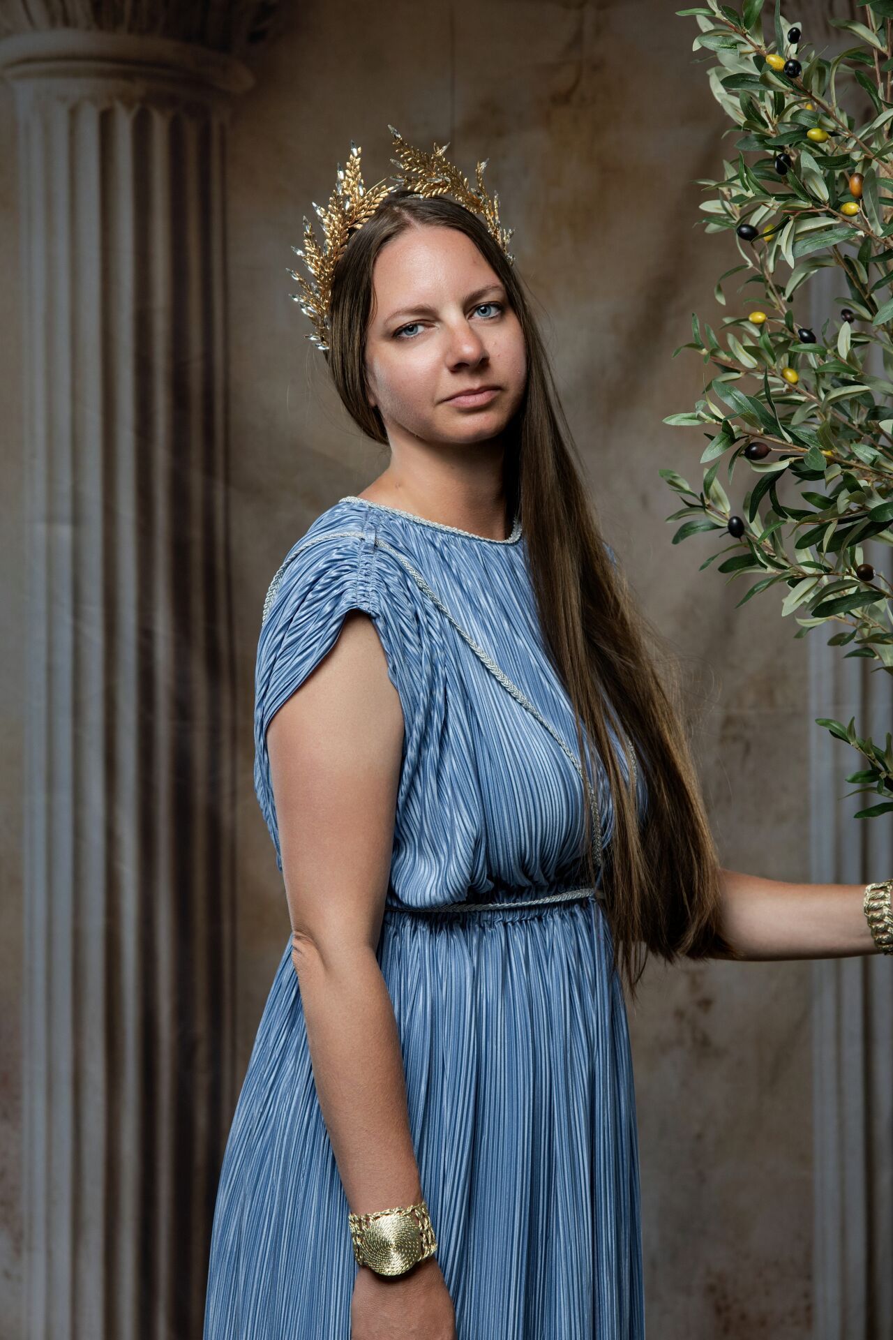 Woman in blue pleated Greek goddess dress with gold crown and bracelet poses by olive tree and marble columns.