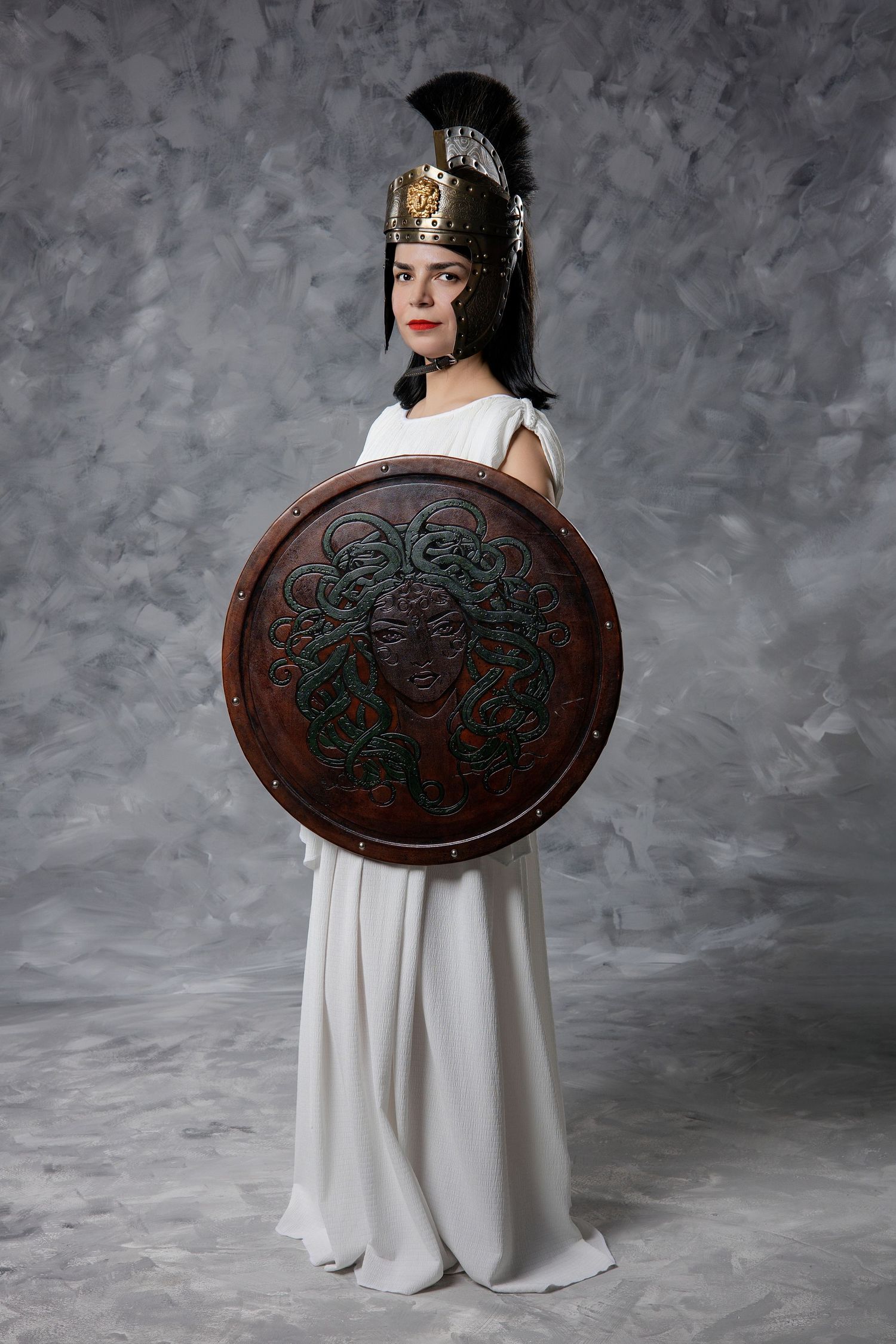 Woman in white Greek dress holding round Medusa shield wearing ornate Corinthian helmet in studio photo.