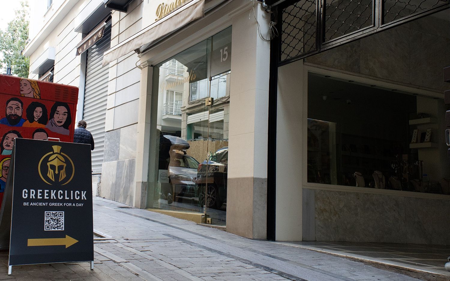 A narrow cobblestone street with white buildings and storefronts on both sides in an urban European setting.
