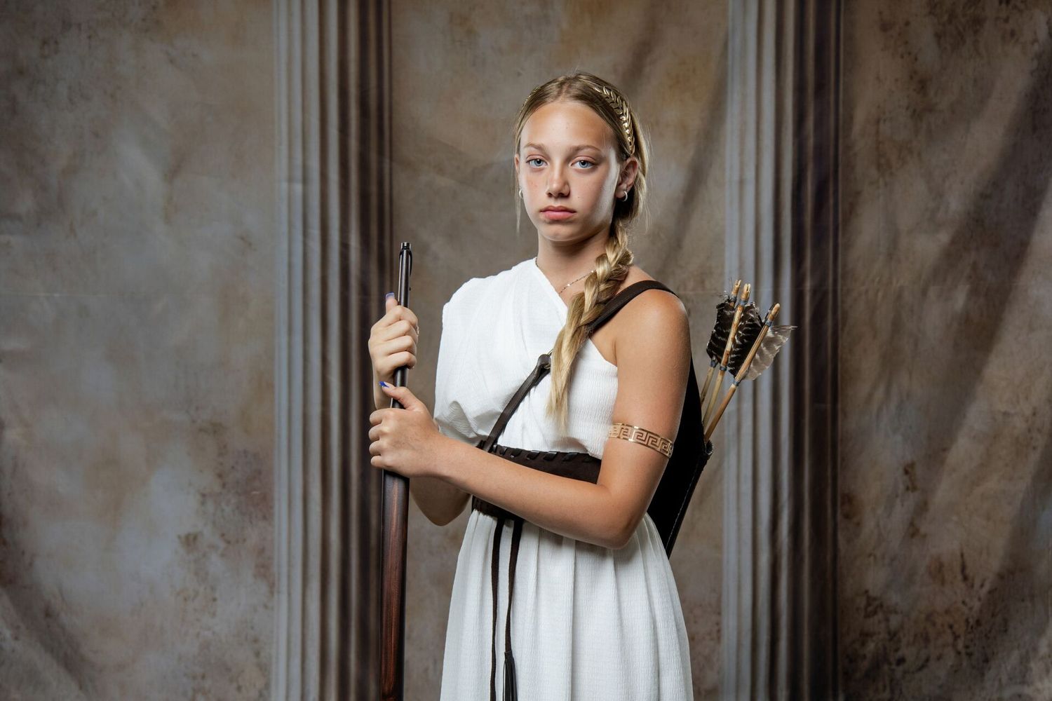 Young woman dressed as Greek goddess Artemis in white dress holding bow and arrows in dramatic studio setting.