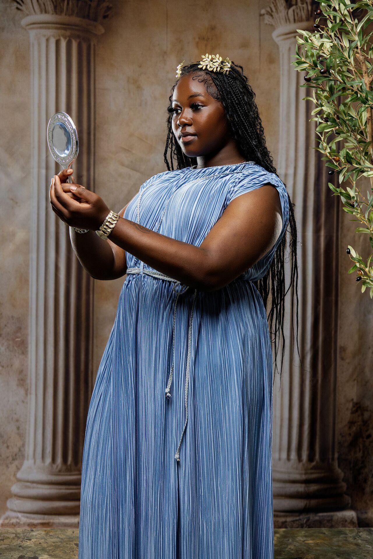 Woman in blue pleated dress holding mirror, gold crown, braids, Greek goddess style portrait.